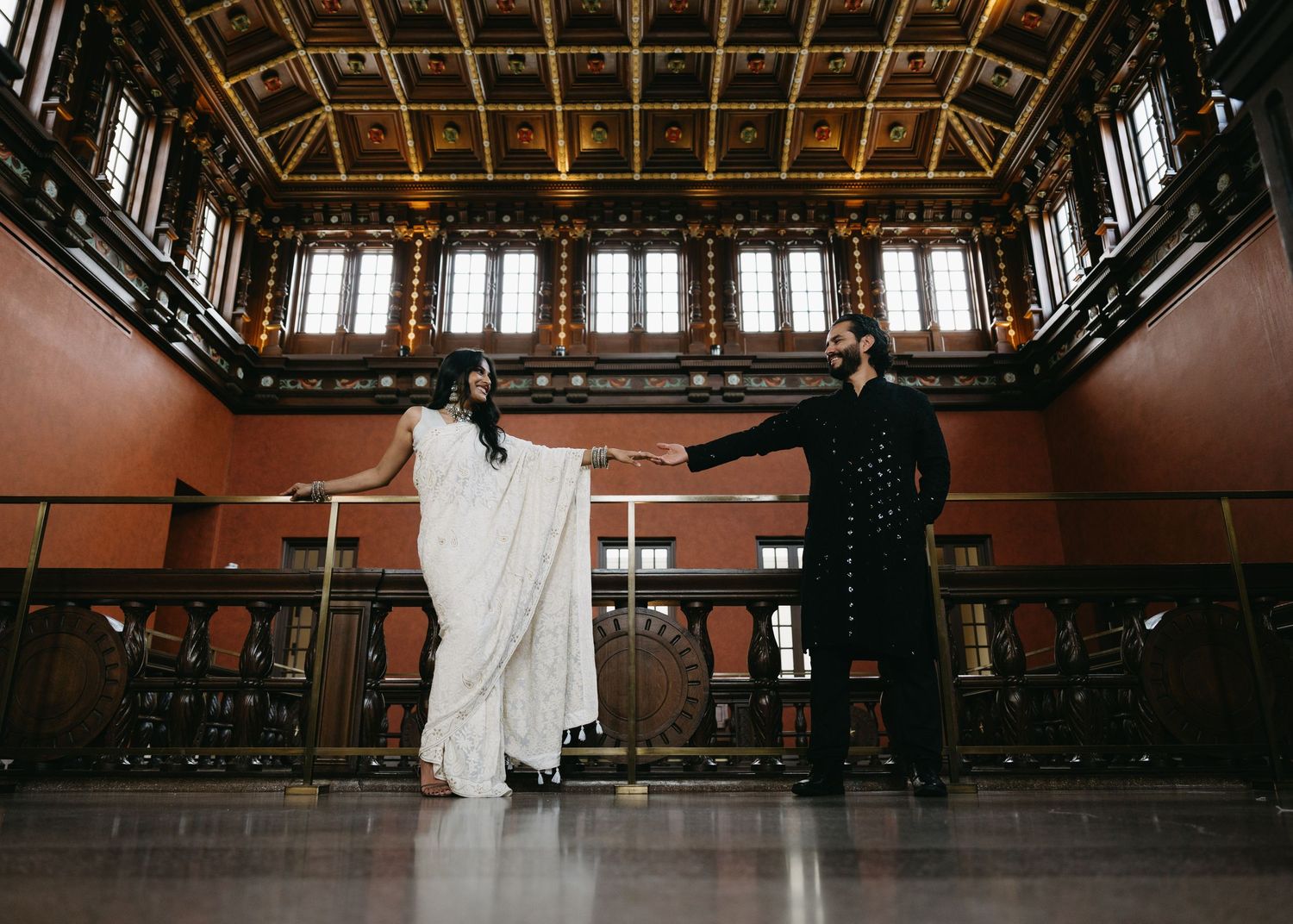 Bride in white saree and groom in dark suit hold hands in an ornate wood-paneled hall during wedding ceremony.