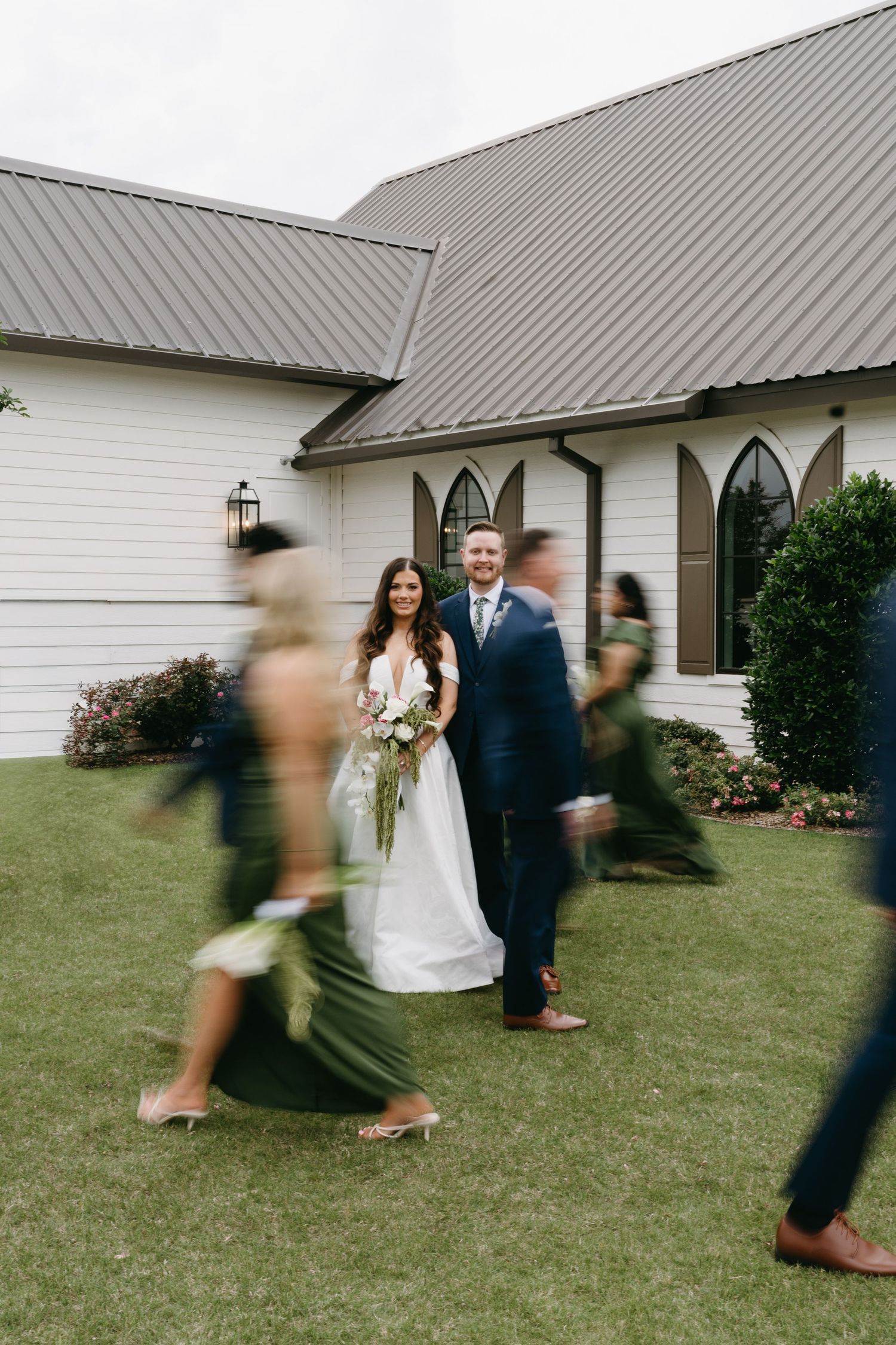 Bride and groom stand smiling outside a white chapel while wedding party members walk past in motion blur.