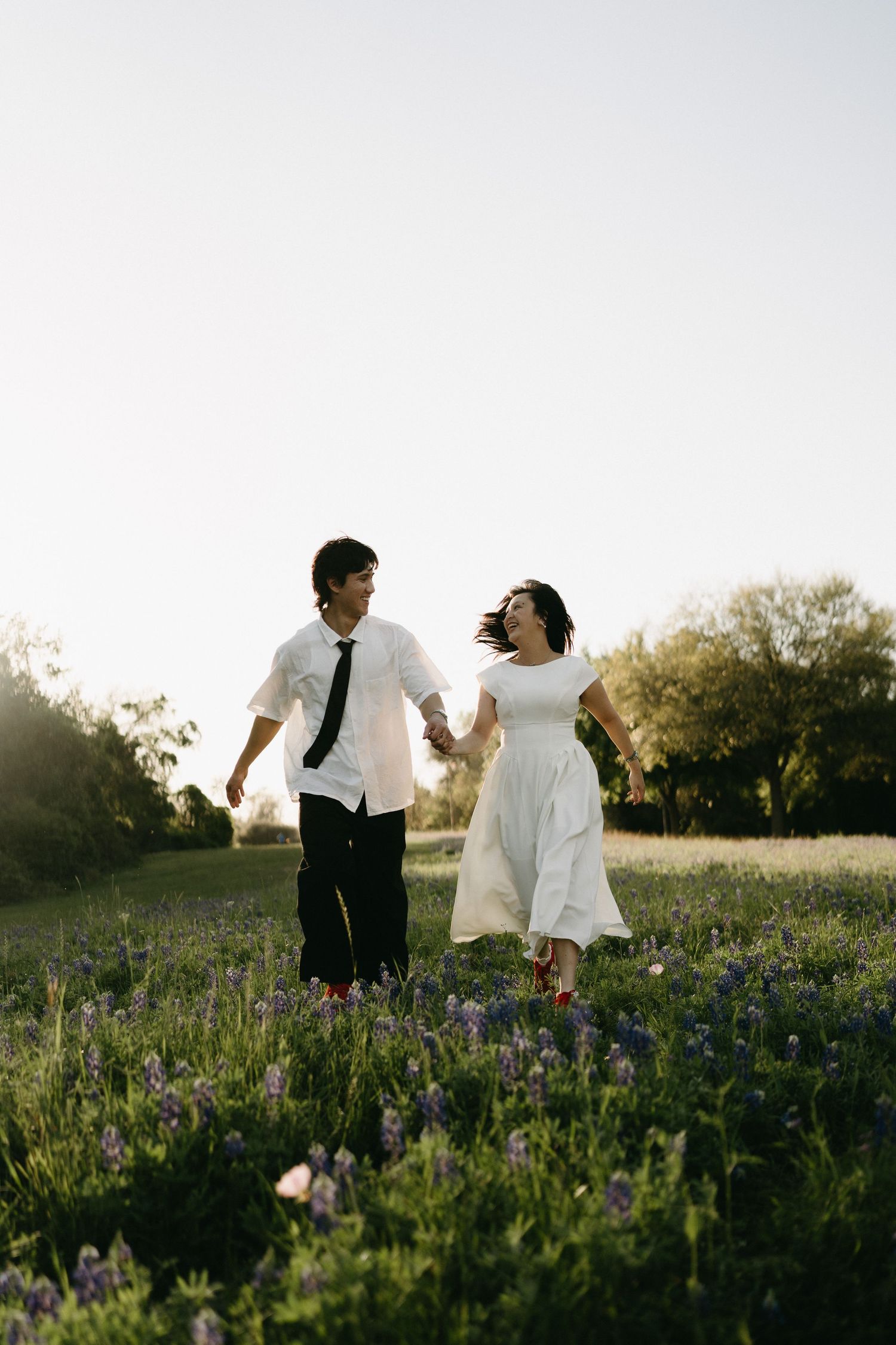 A couple runs joyfully through a wildflower meadow at sunset, with her in a white dress and him in a white shirt and tie.