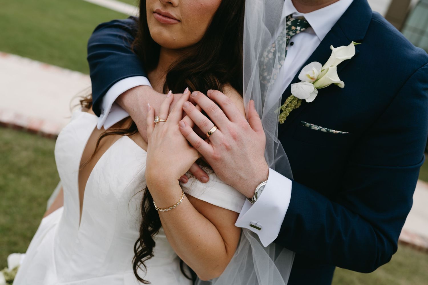 Groom in navy suit embracing bride in white dress, showcasing wedding rings on clasped hands outdoors.