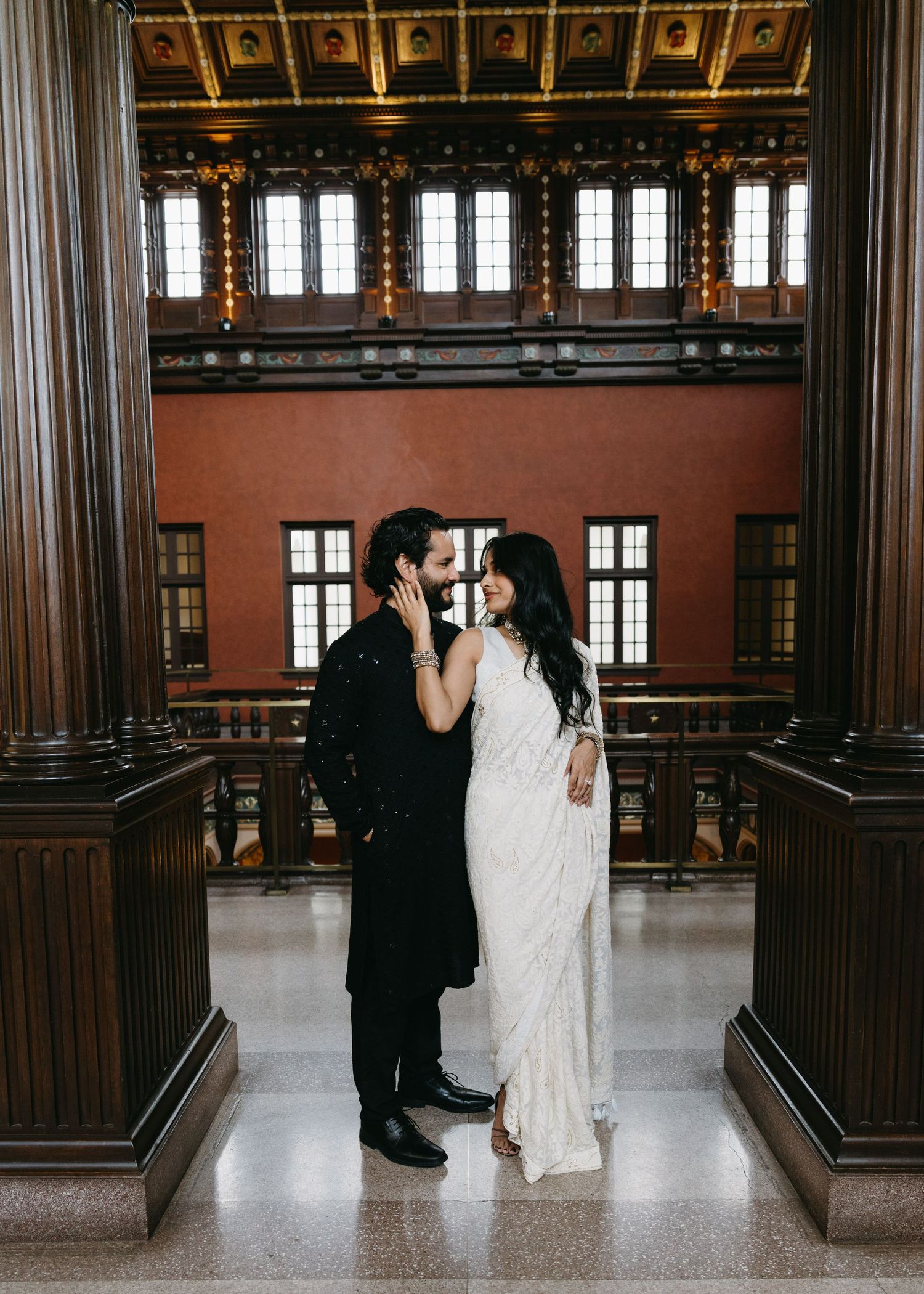 Couple in wedding attire embrace in elegant courthouse interior with tall columns and ornate architecture.