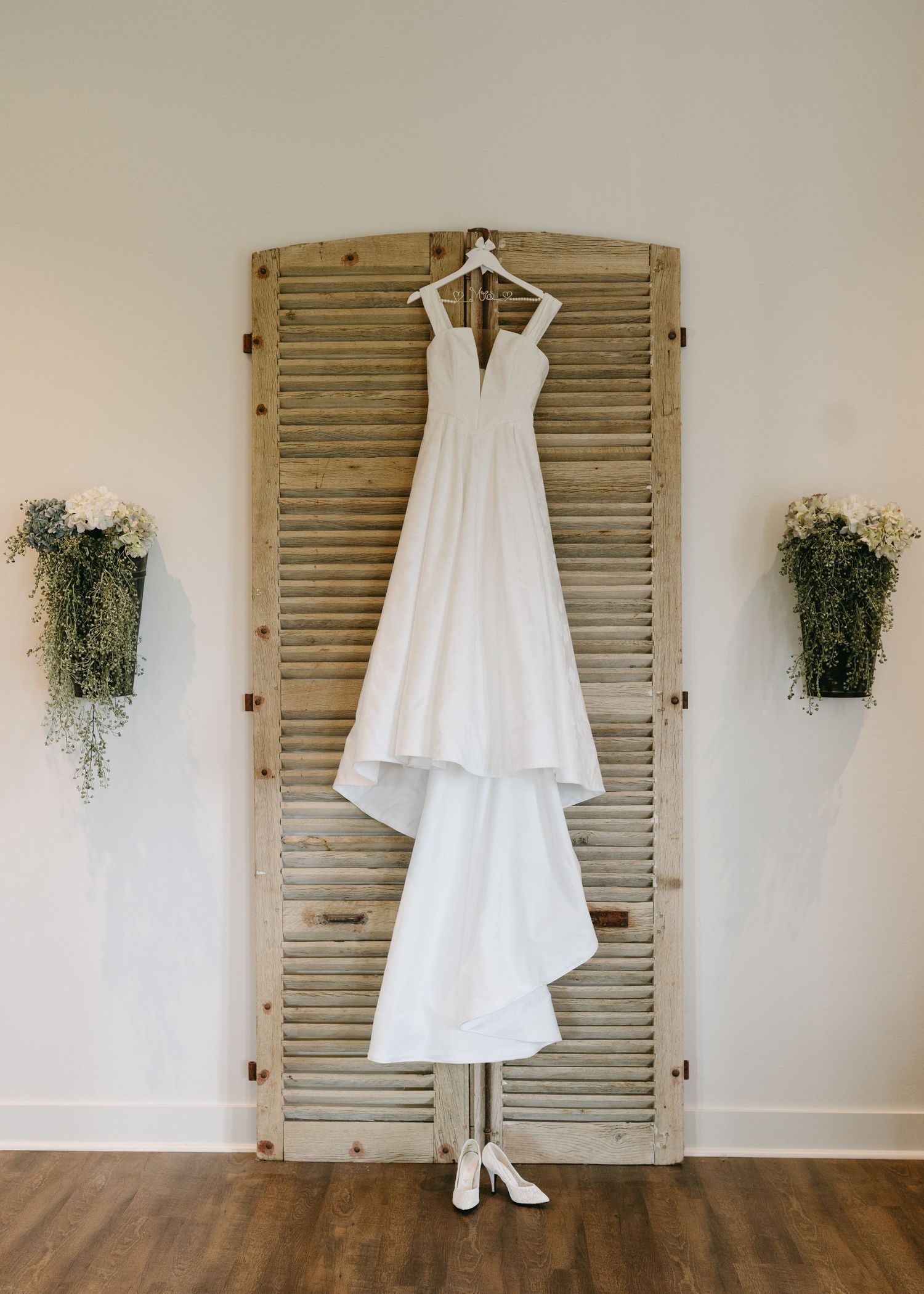 Minimalist white A-line wedding dress hanging on a rustic wooden shutter with white heels and floral wall decor.