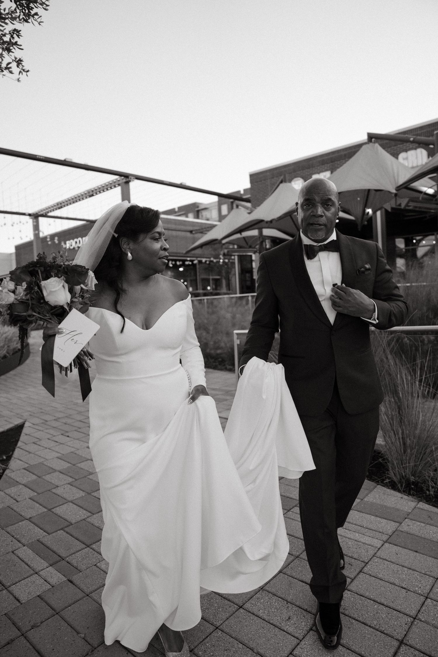 A couple walks together on a paved patio area while holding umbrellas in formal wedding attire.