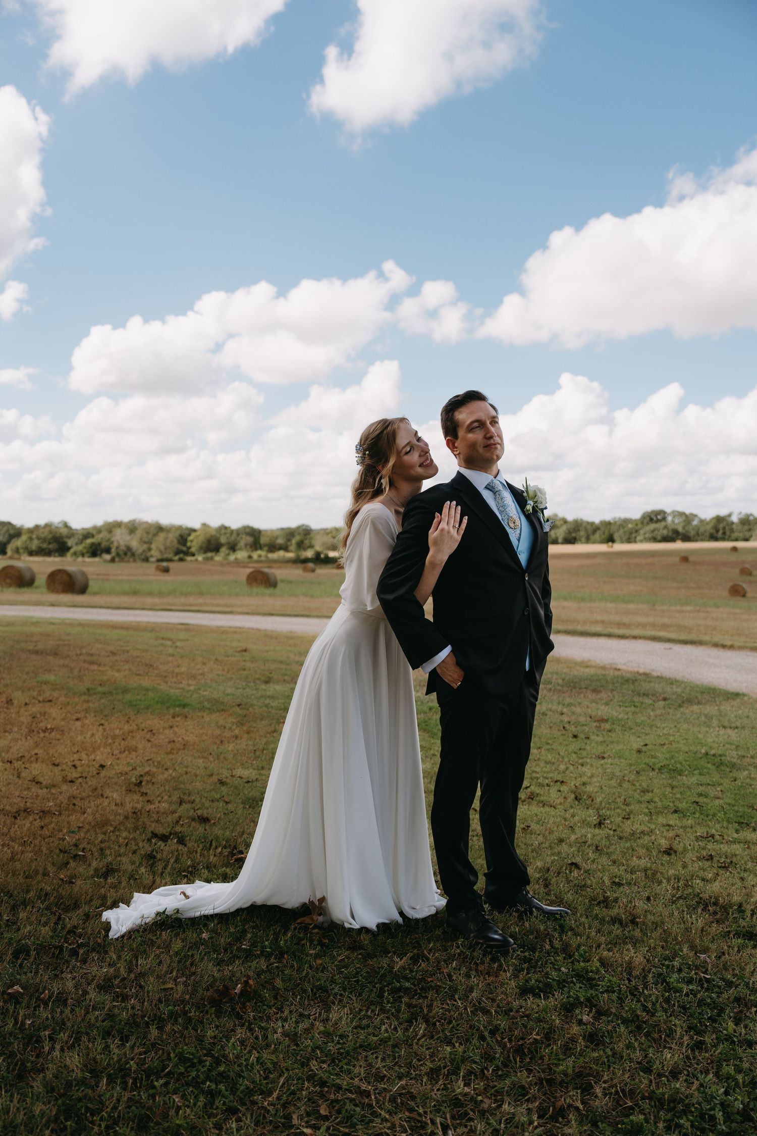 A couple shares a romantic moment in their wedding attire while standing in a scenic field with hay bales and blue skies.