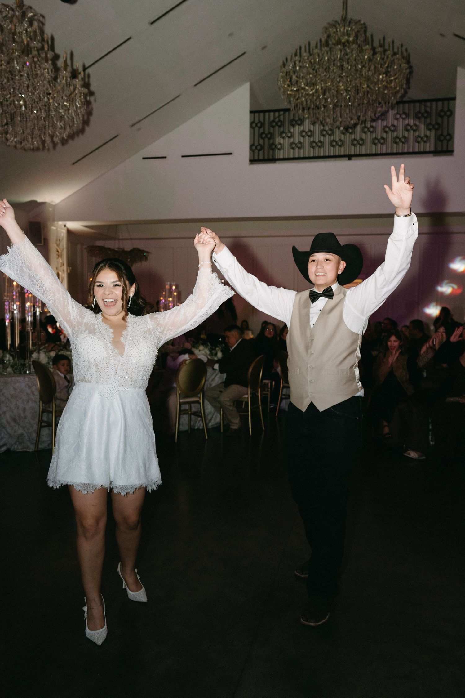 Wedding party dancing energetically under ornate chandeliers in a dimly lit reception venue.
