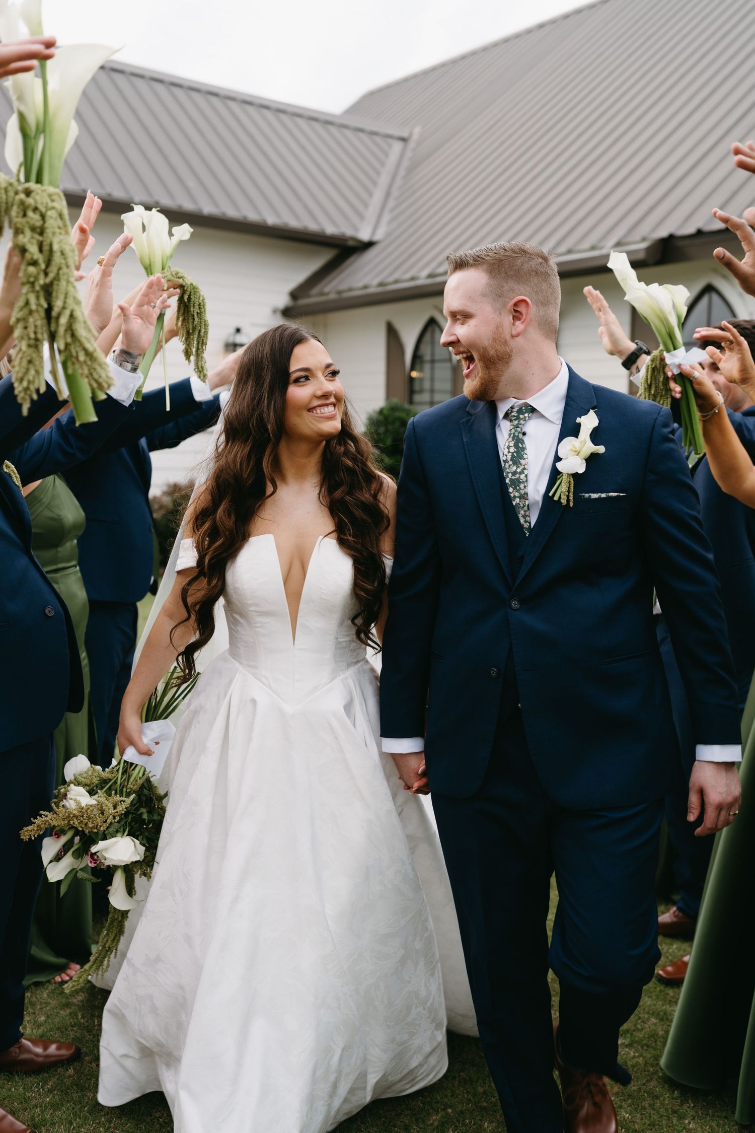 A smiling bride and groom walk hand-in-hand through a floral arch of calla lilies at their outdoor wedding ceremony.