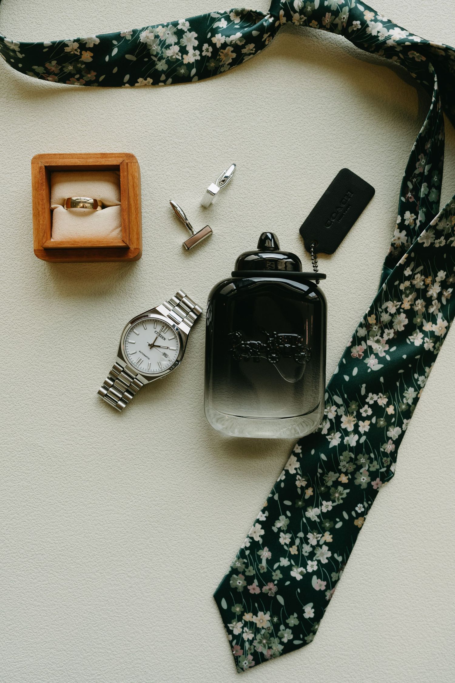 Groom's wedding accessories flat lay featuring a floral tie, silver watch, gold ring, cufflinks, cologne, and Coach keychain.