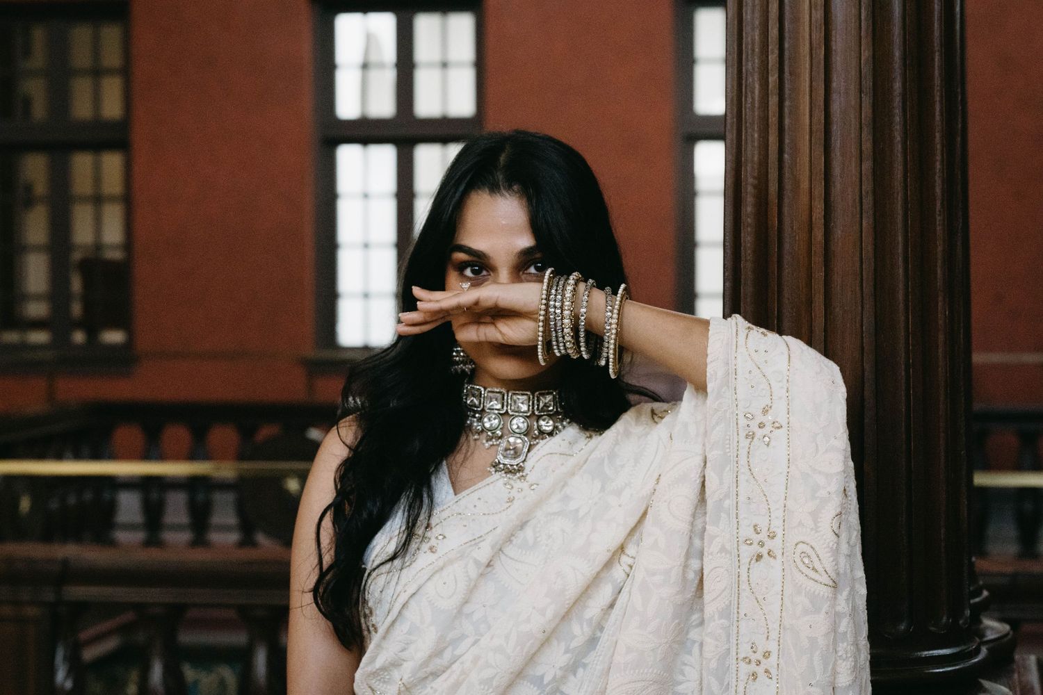 Woman in white embroidered saree and silver jewelry poses gracefully against a brick column.