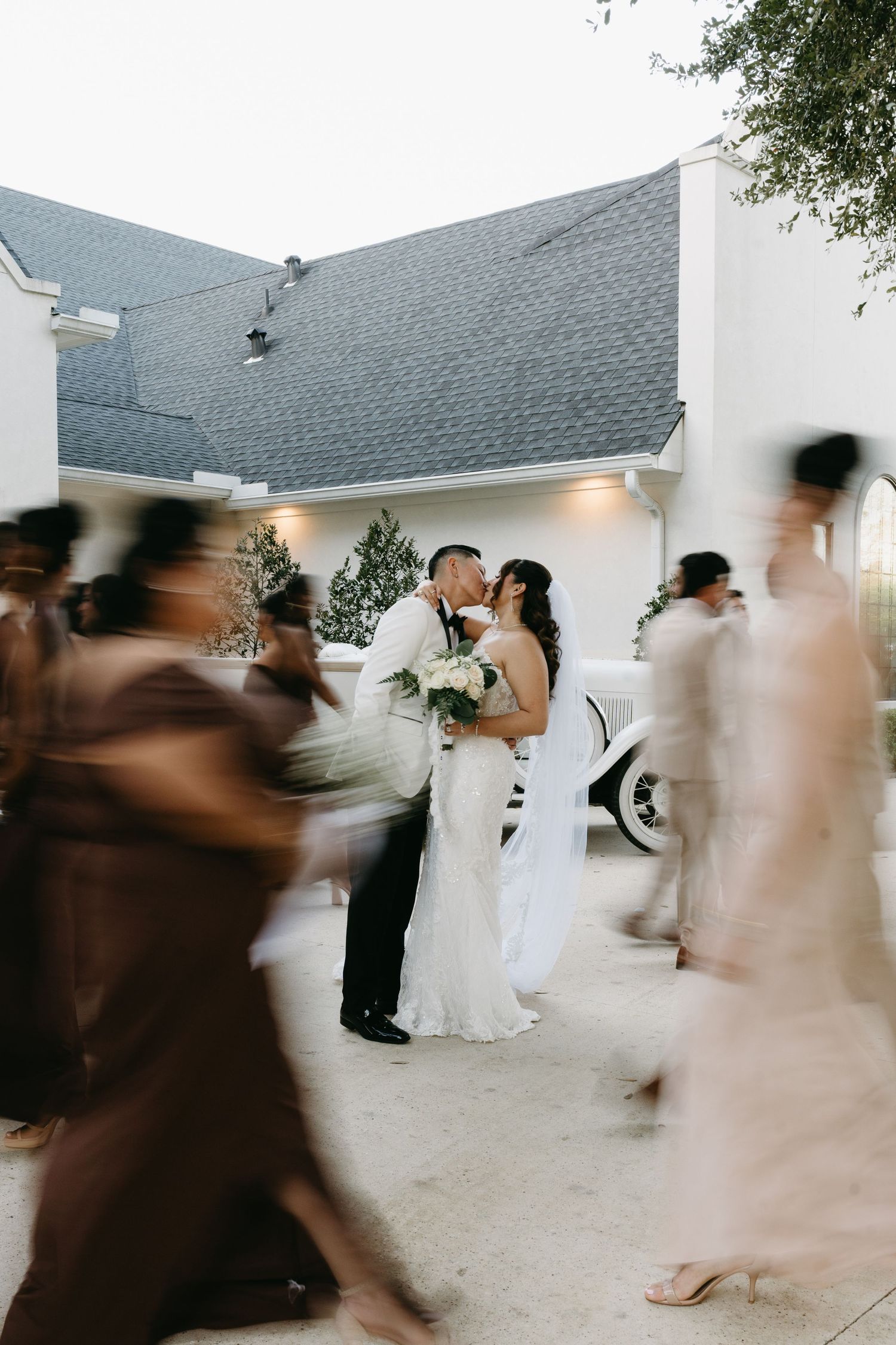 Motion blur captures wedding guests dancing at an outdoor reception with white walls and gray roofing in the background.