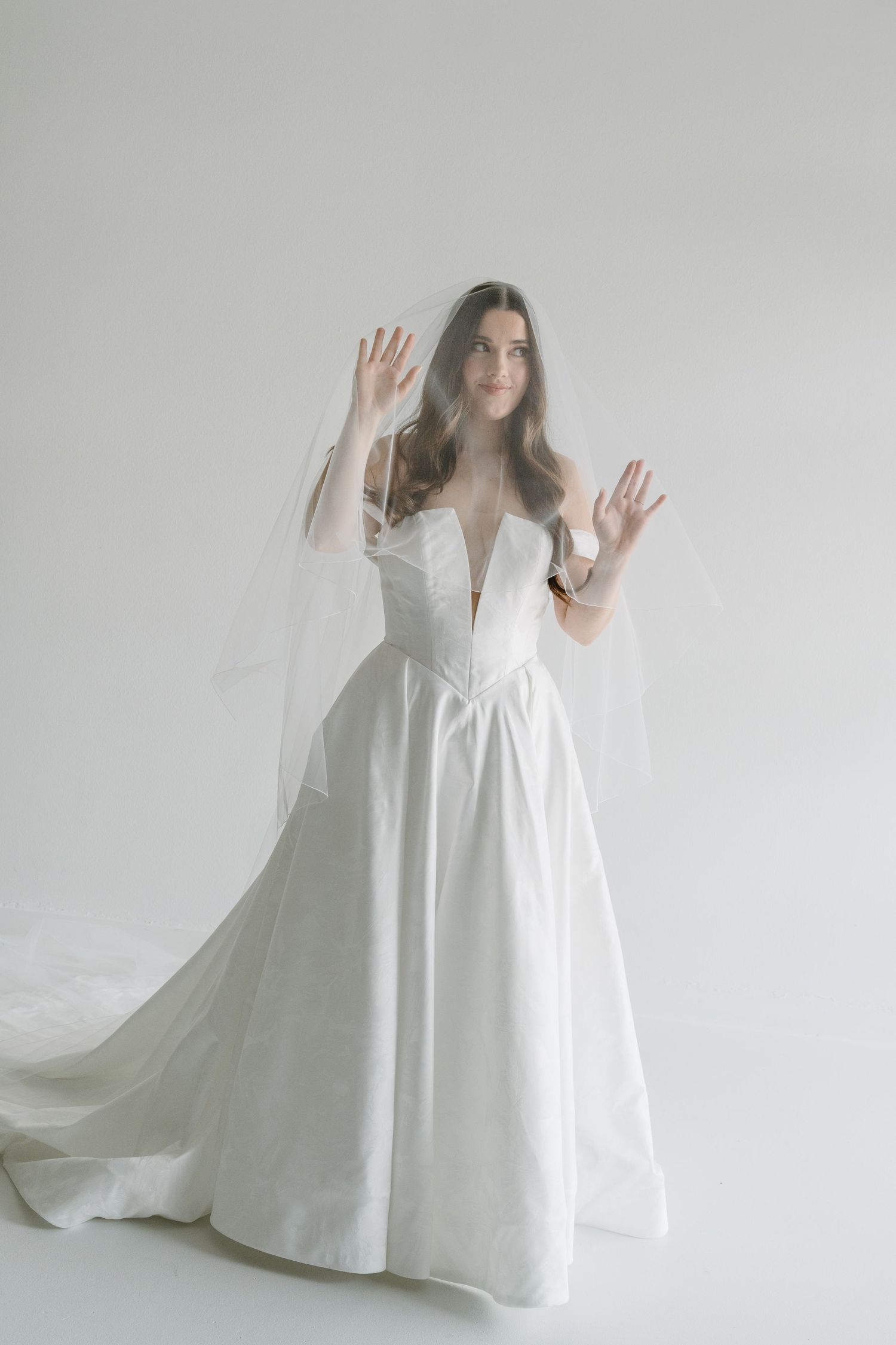 Bride in white ball gown with deep V-neckline and flowing veil raised over hands in bright studio.