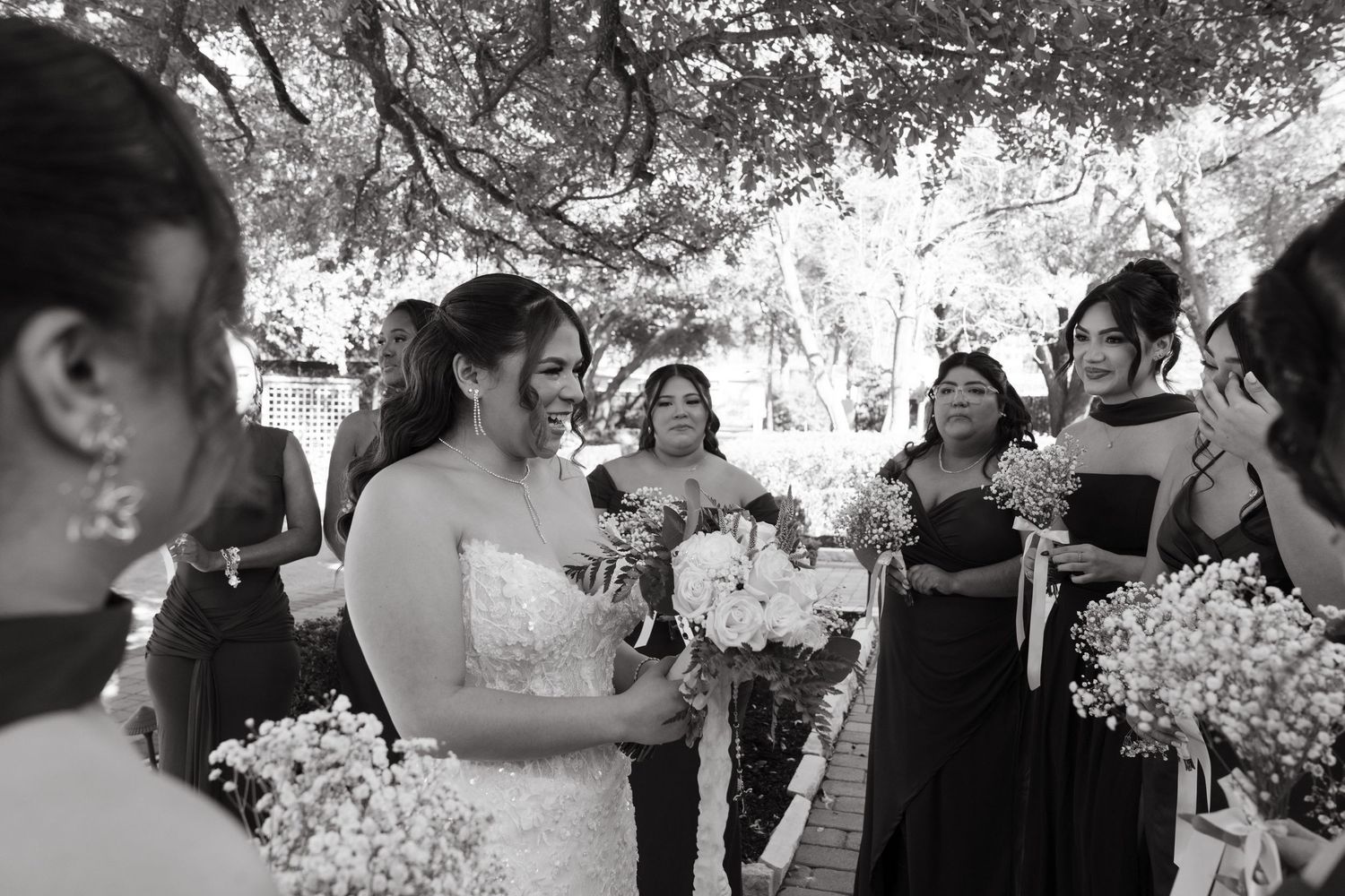 A black and white photo of a bridal party laughing together while holding bouquets outdoors under trees.