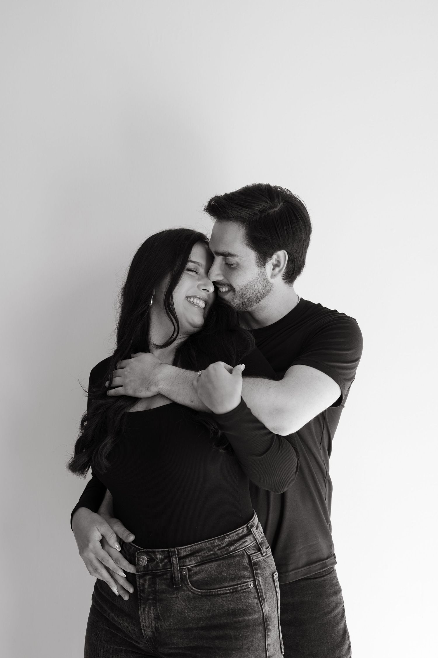 A couple in black clothing embraces intimately in a series of romantic black and white photographs against a white wall.