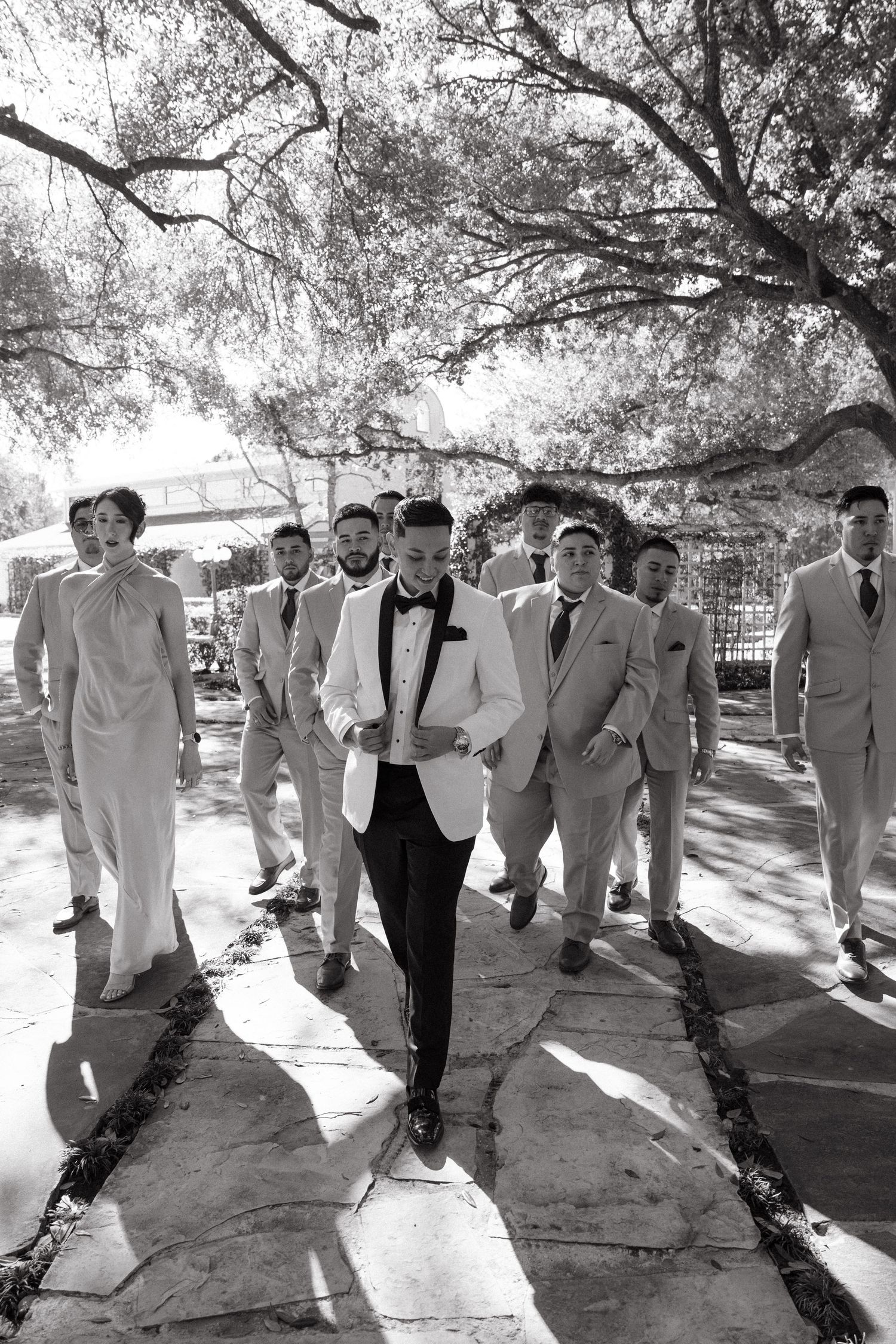 A group of groomsmen in light suits and dark bowties walk together in a line under shady trees on a sunny day.