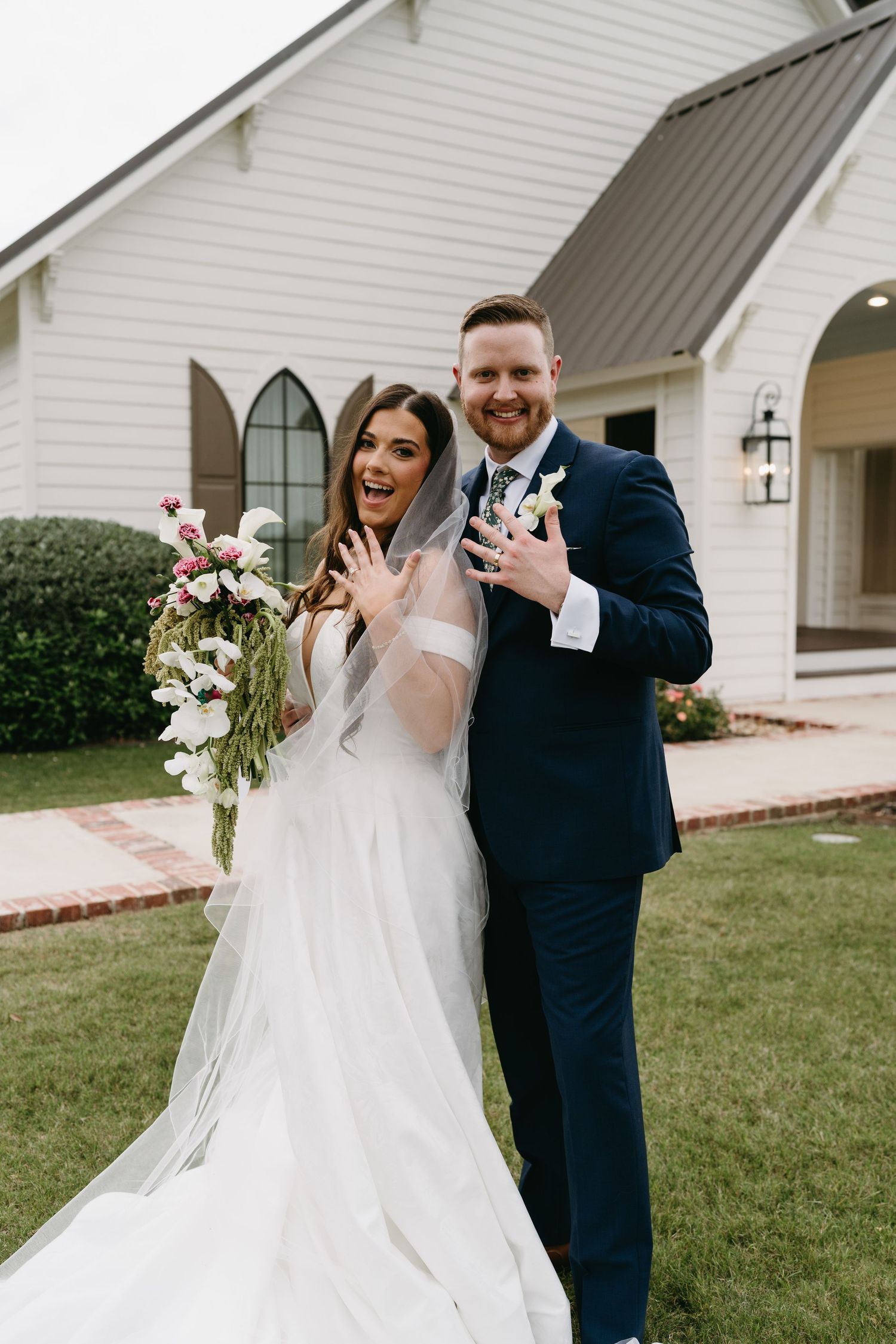 Joyful newlyweds pose outside white chapel, bride in tulle gown showing ring, groom in navy suit holding champagne.