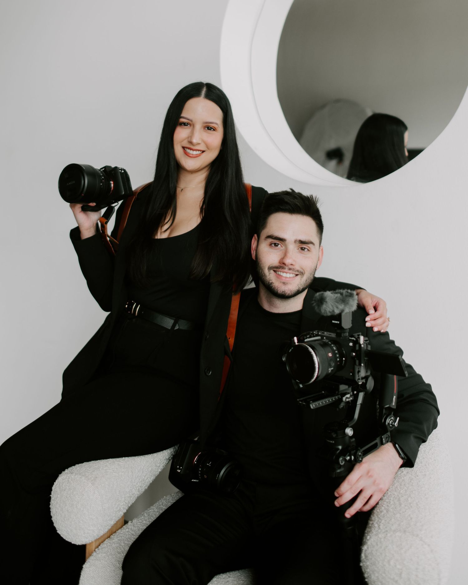 Two photographers in black outfits pose with professional cameras against a white wall with a circular mirror.