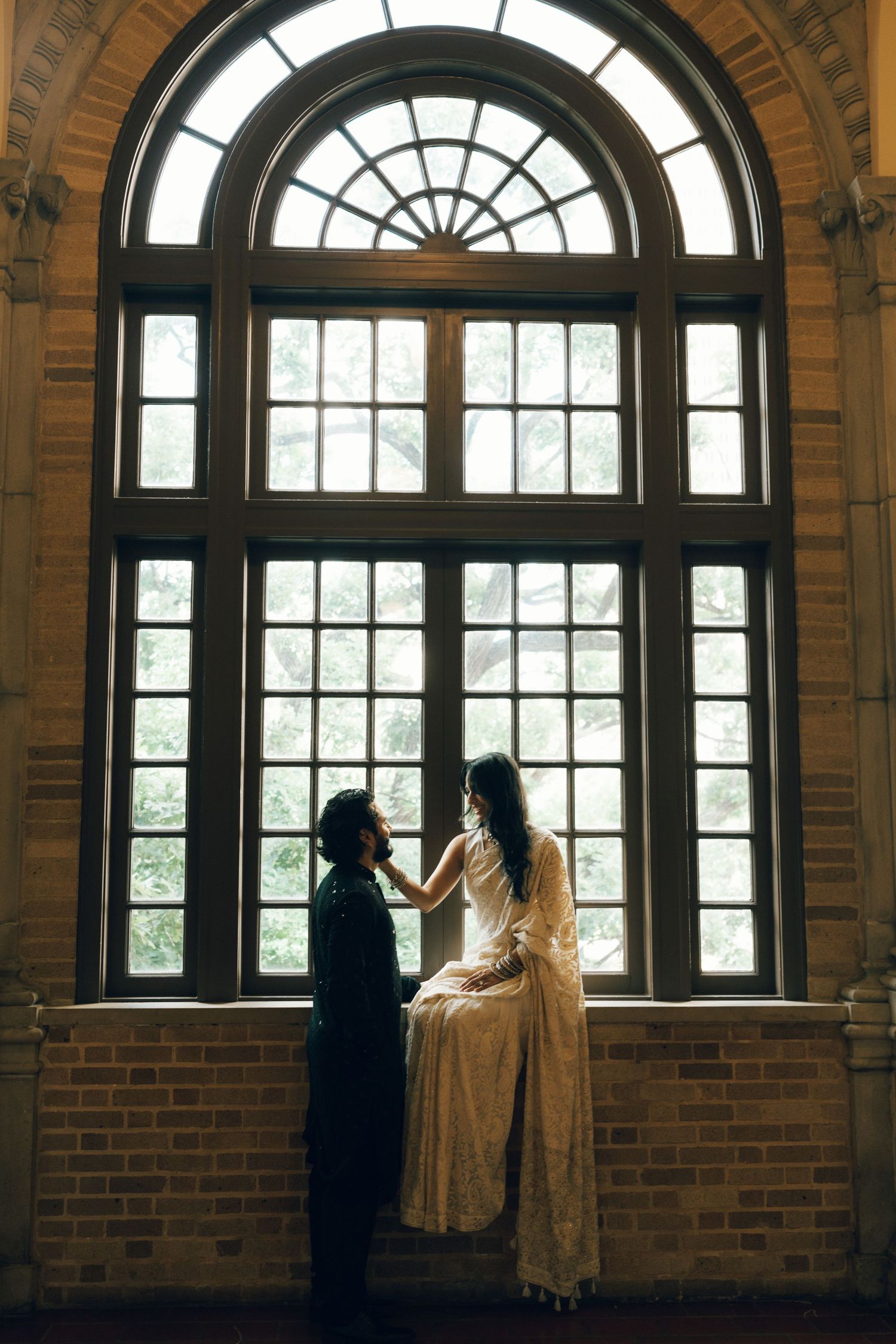 Couple in elegant attire sharing a romantic moment by a grand arched window in a historic brick building.