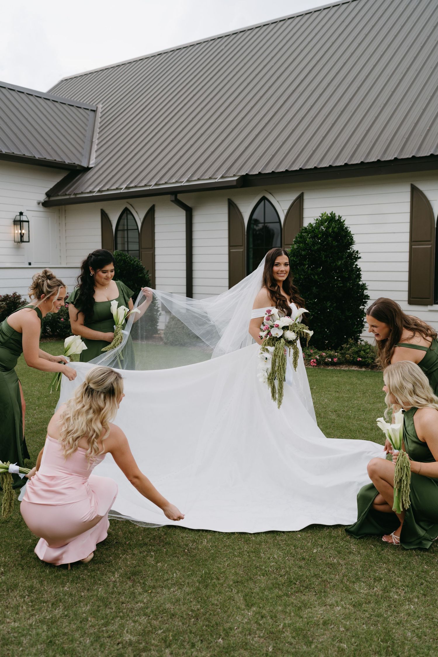 Bride in white gown and veil surrounded by bridesmaids in green dresses outside a white chapel.