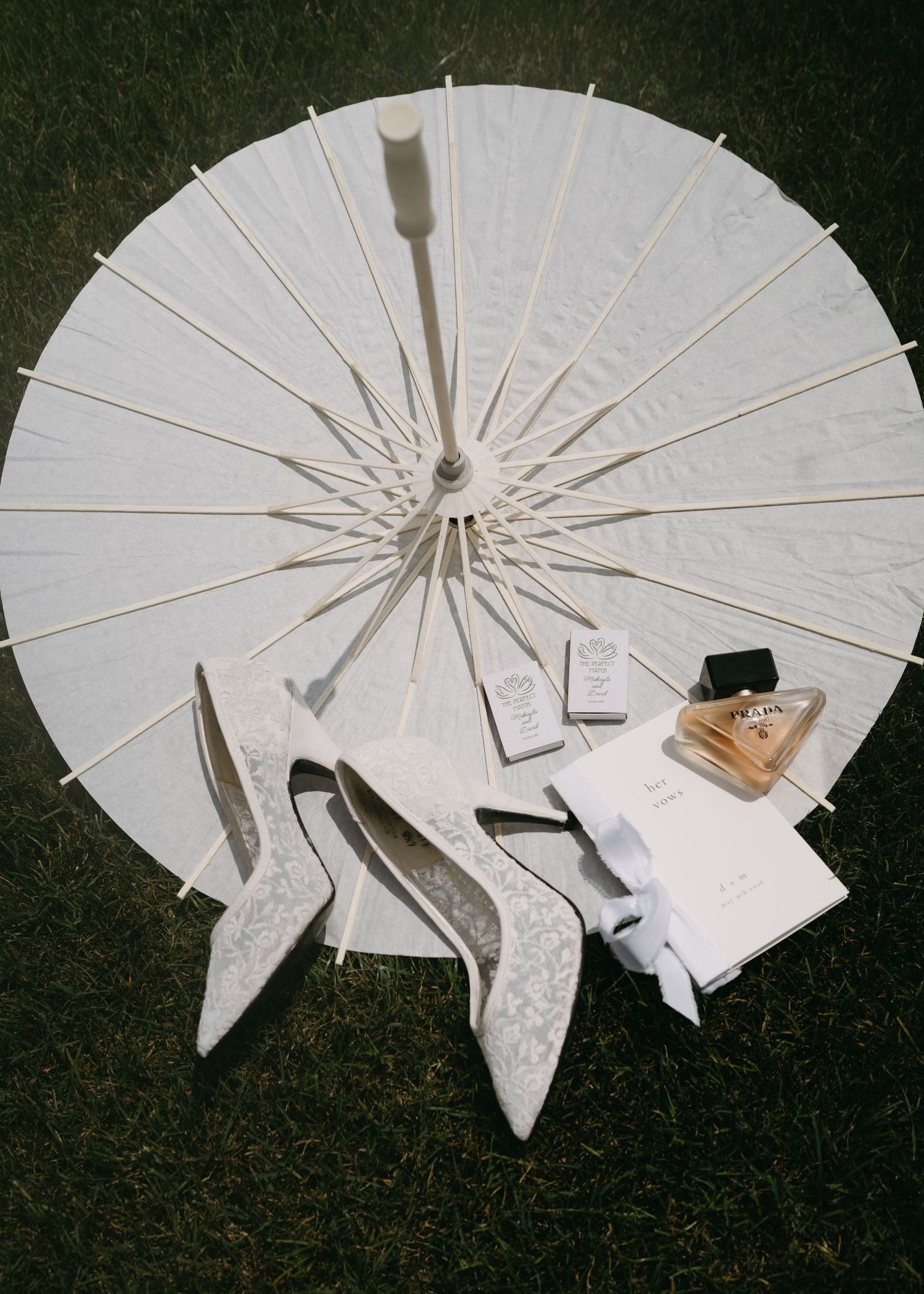 White parasol umbrella with lace bridal heels, perfume bottle, and wedding stationery arranged on green grass.