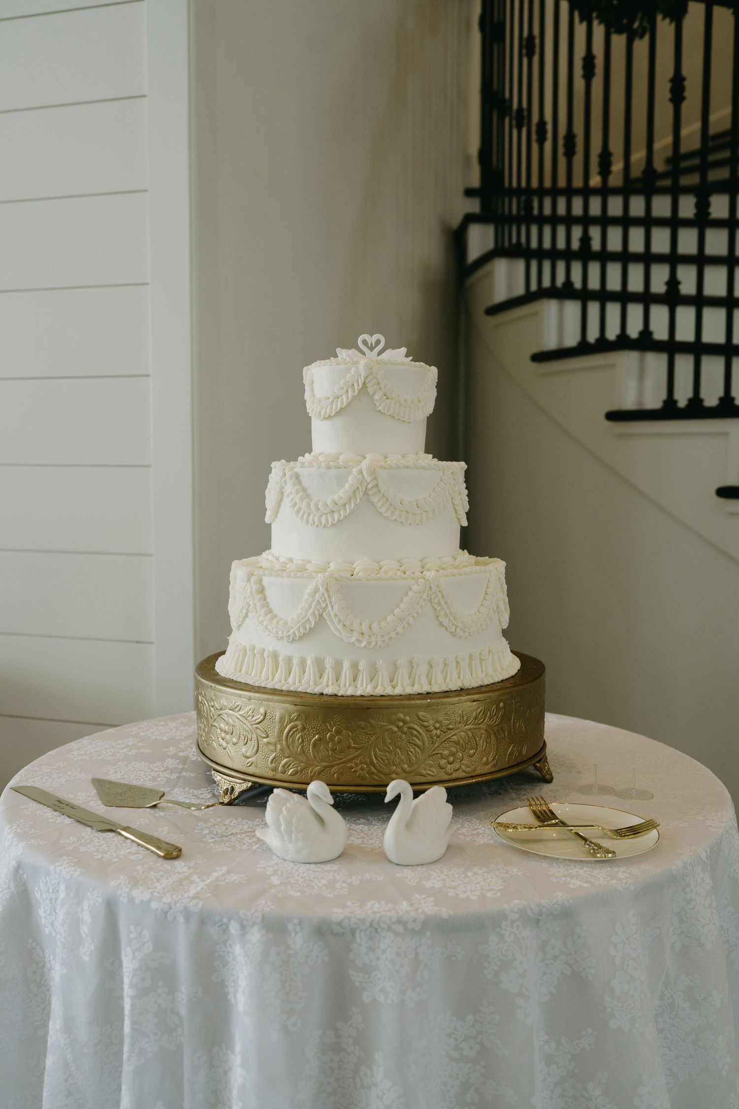 Elegant four-tier white wedding cake with swan topper on gold stand, decorated with swag frosting details and two swan figurines.