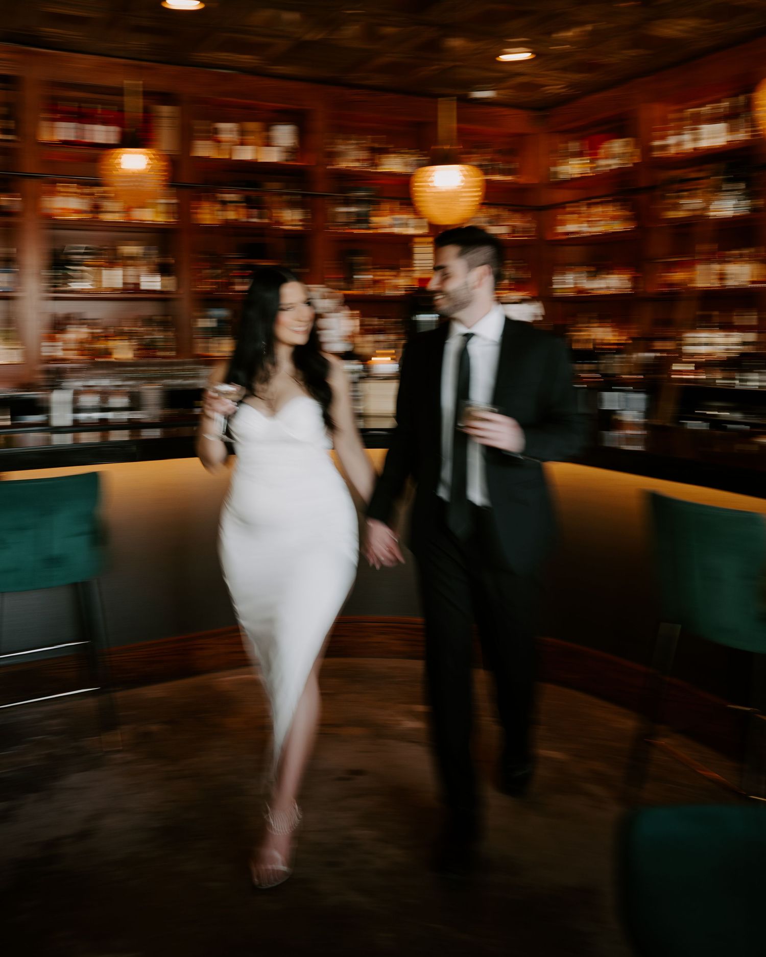 A couple walks together in a cozy bar with wood-paneled walls, warm lighting and emerald green booth seating.