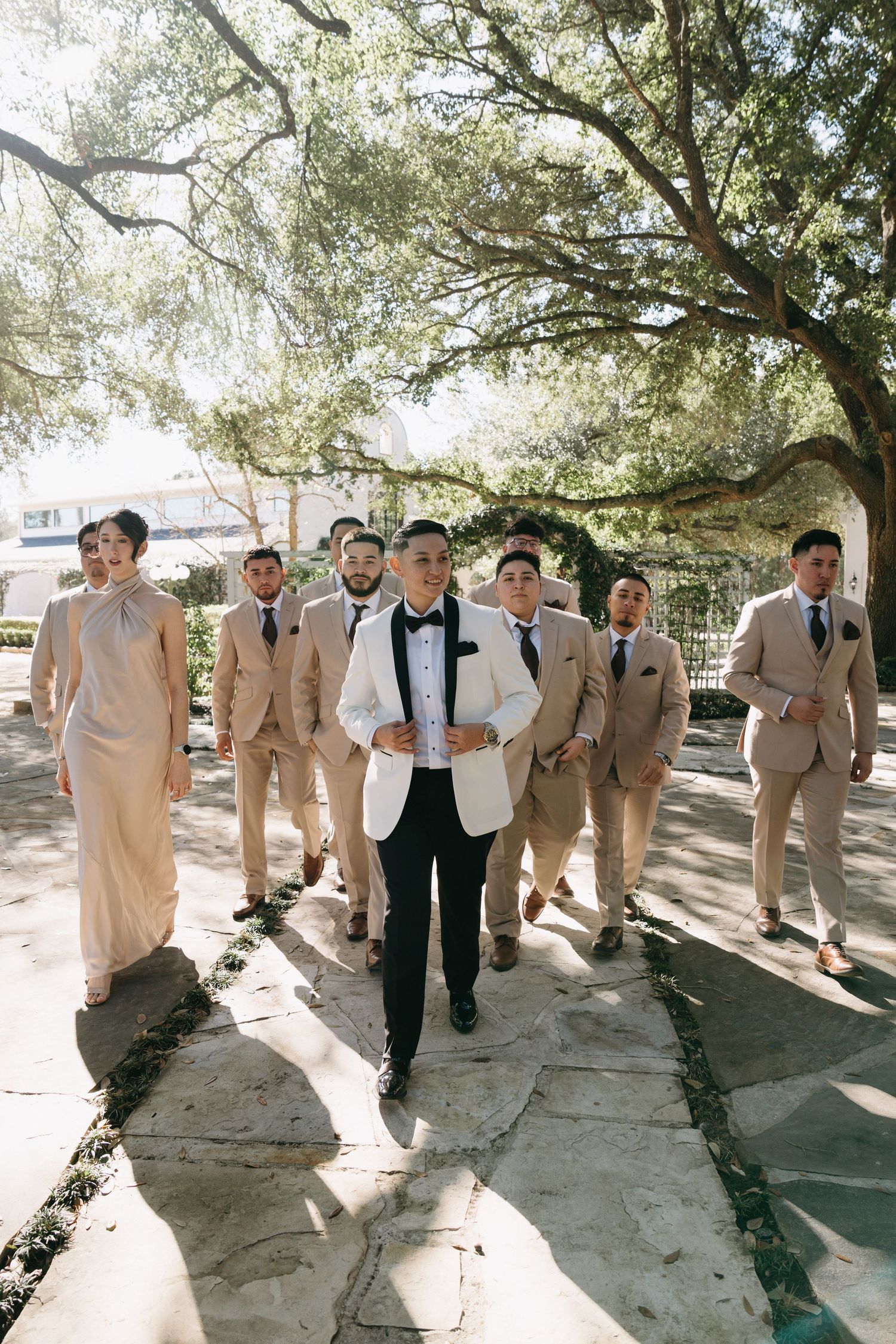 A wedding party in light beige suits and dark ties walks together in a line under shady trees on a sunny day.