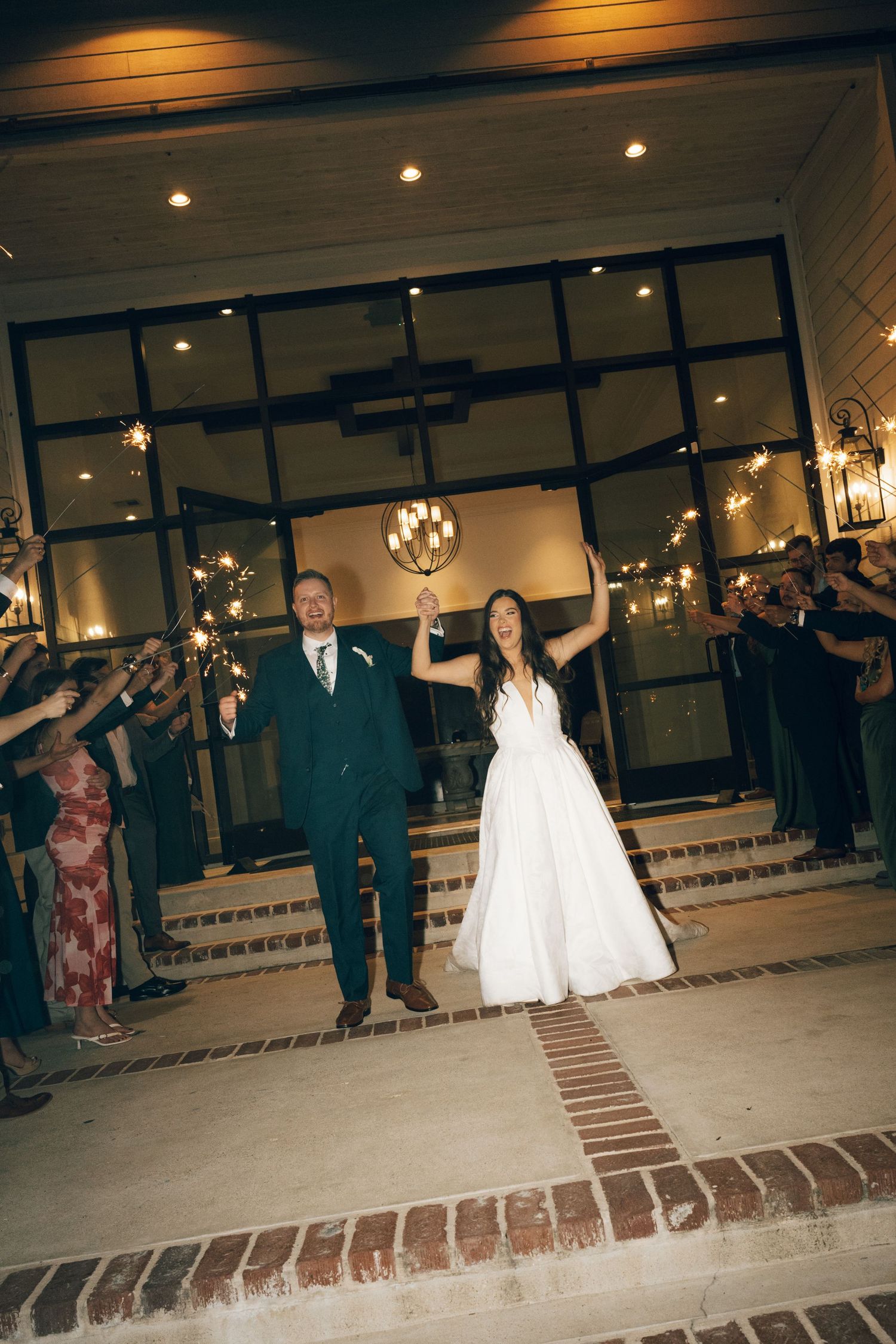 Bride and groom exit wedding venue through sparkler send-off, raising hands in celebration at night.