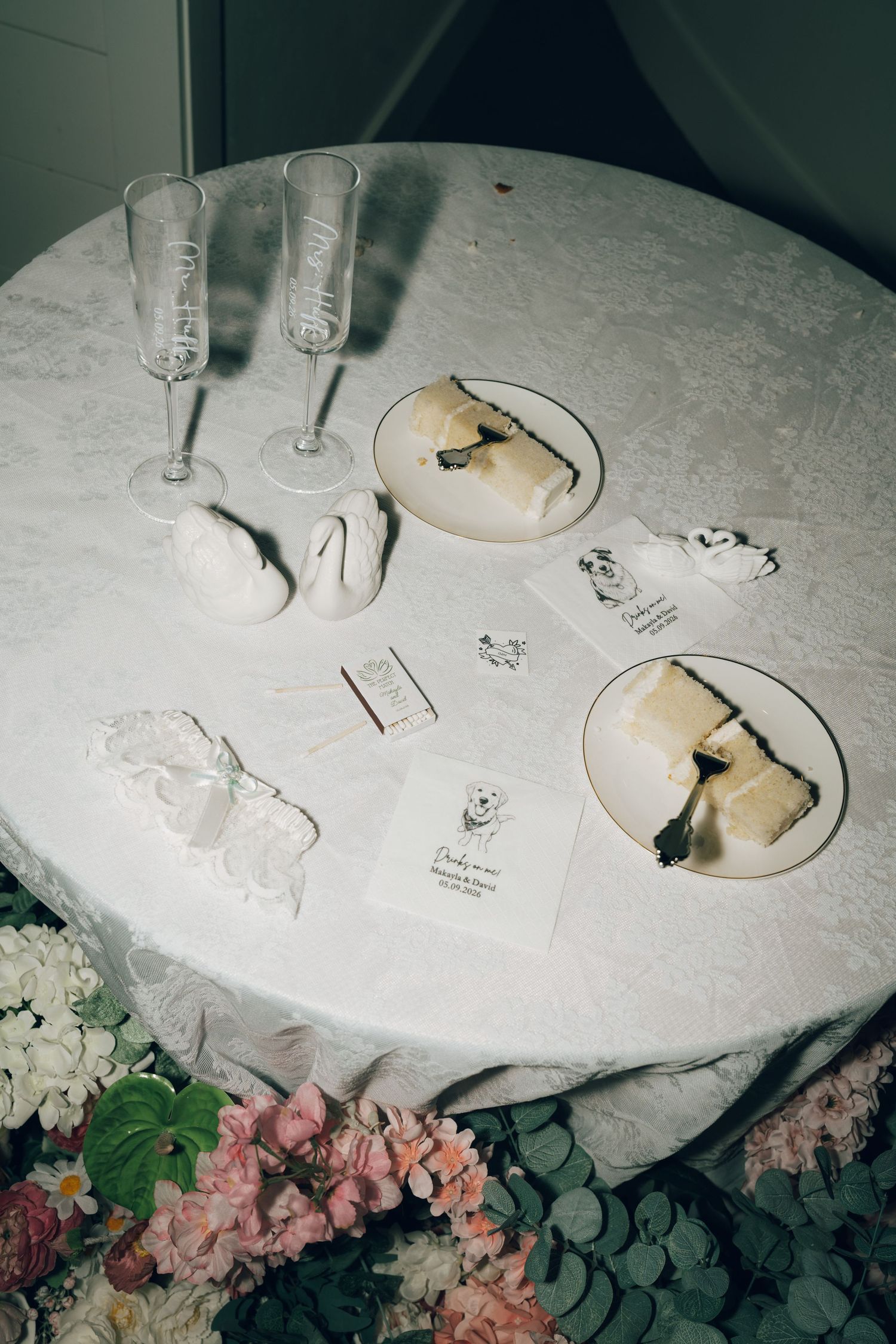 Elegant wedding table with champagne flutes, swan figurines, cake slices on plates, and floral arrangements below.
