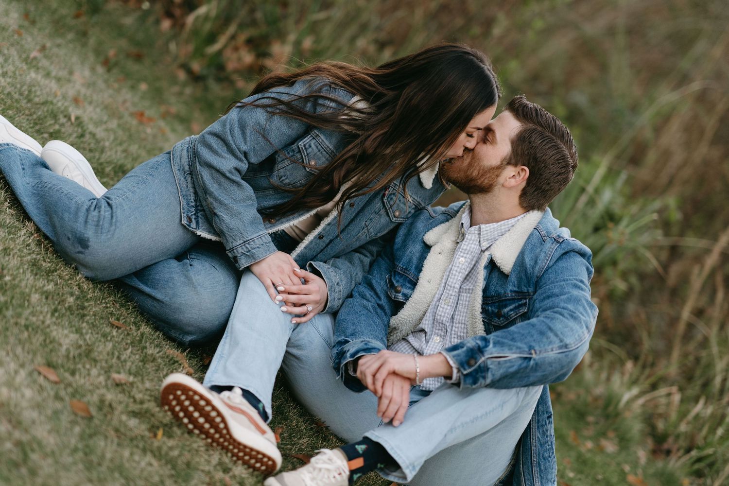Couple sitting in casual denim outfits embracing on grassy ground during outdoor photoshoot.