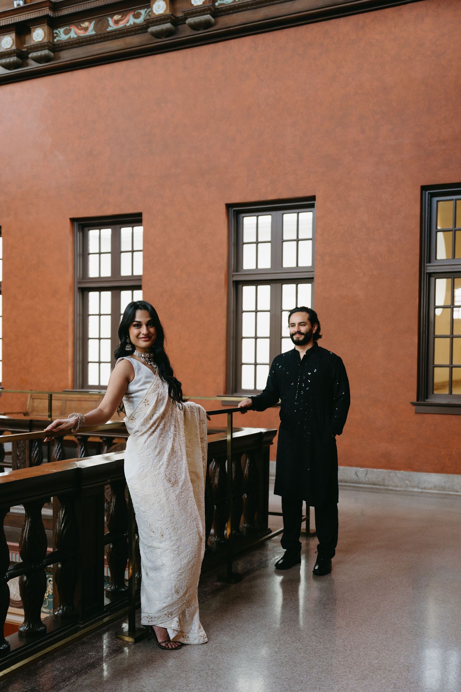 Indian couple in traditional attire posing elegantly in a grand hall with warm terracotta walls and wooden railings.