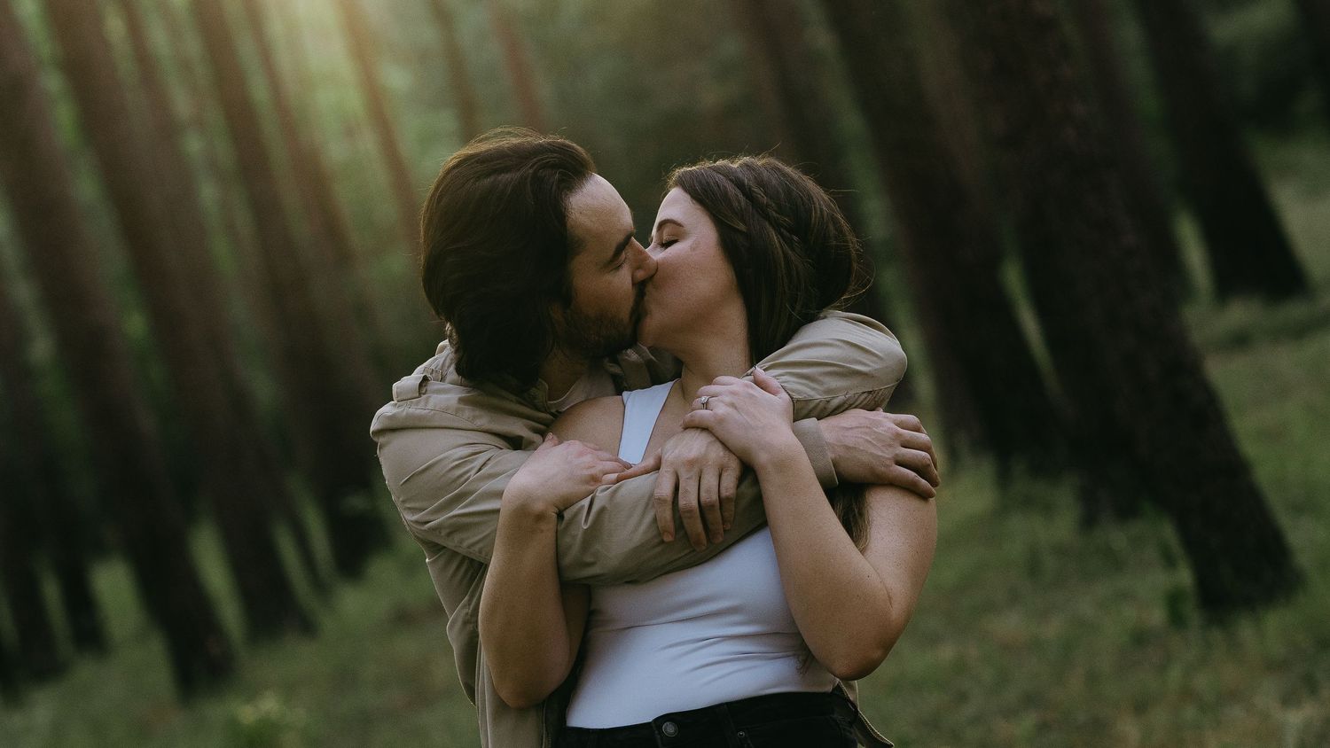 A couple shares a romantic kiss while embracing in a lush green forest with tall trees in the background.