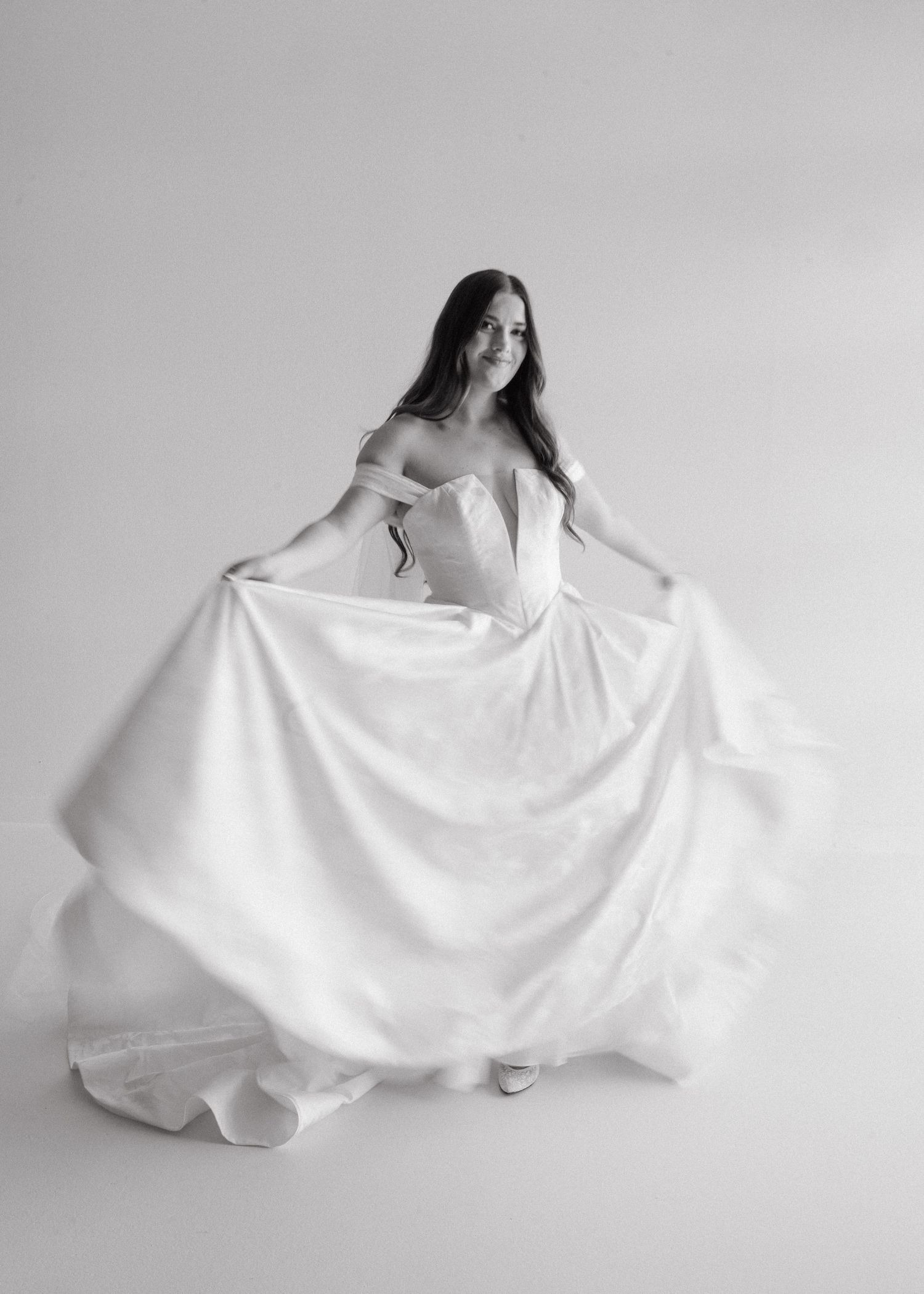 Black and white photo of a bride in an elegant off-shoulder ball gown, gracefully spreading her skirt in a studio.