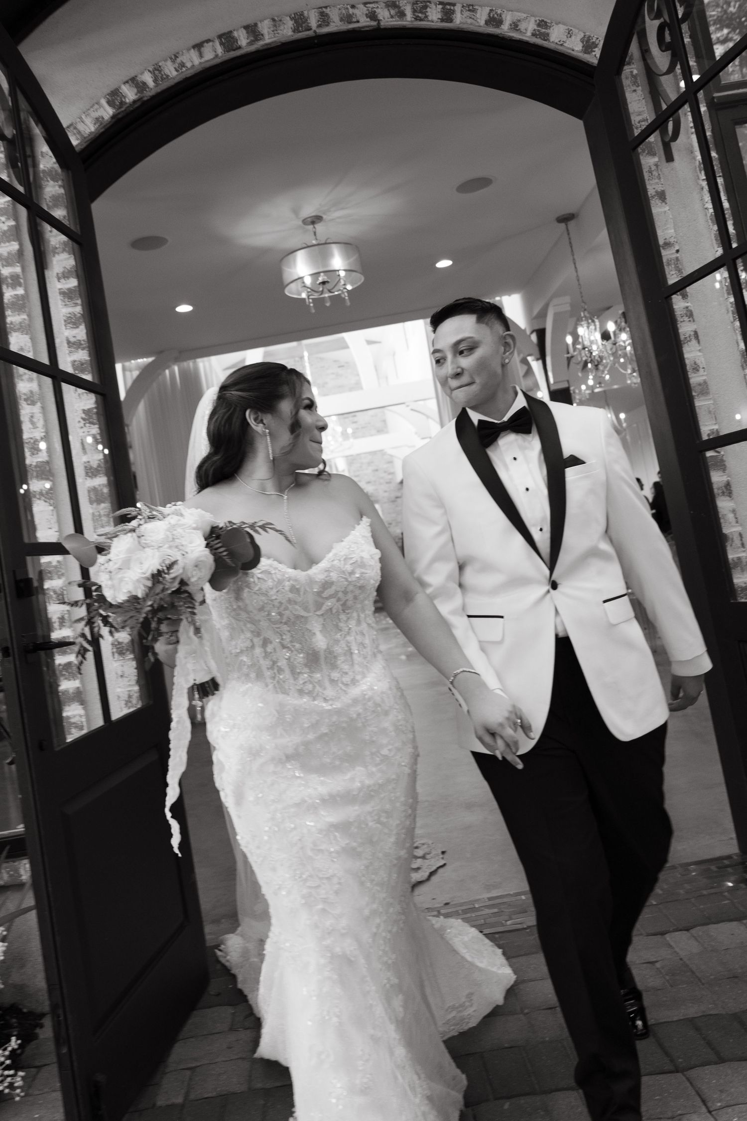 Black and white photo of a bride and groom holding hands, walking through elegant venue doors, bride in lace gown, groom in white tuxedo.