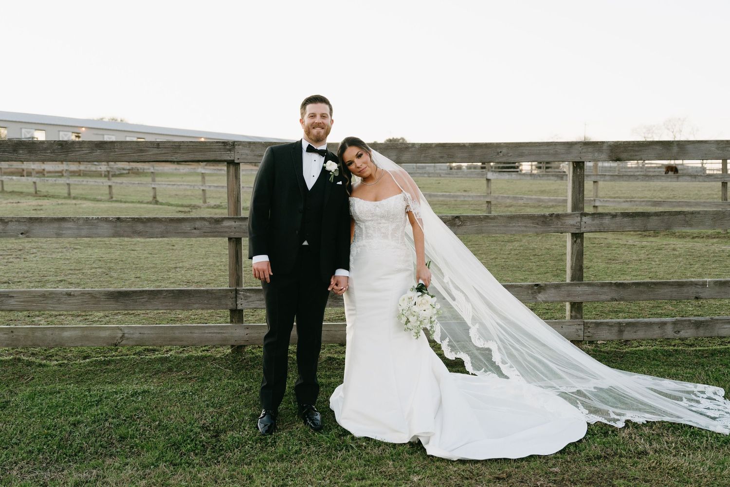 Bride in white gown with veil and groom in black tuxedo posing together by a wooden fence on a farm.