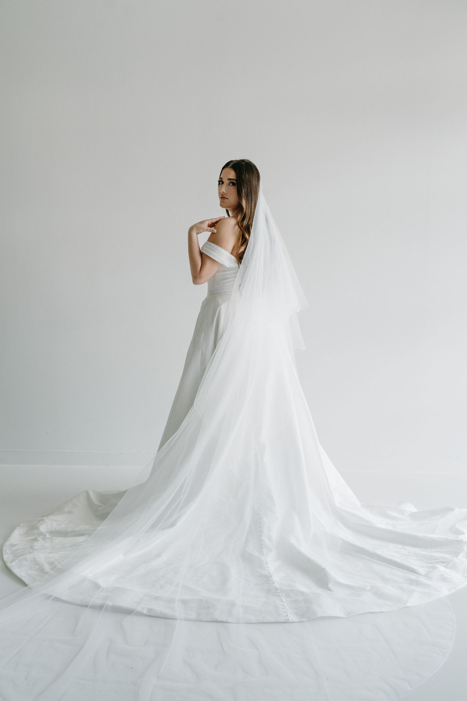 Bride in off-shoulder white wedding gown with long cathedral veil and train, posing gracefully against a white background.