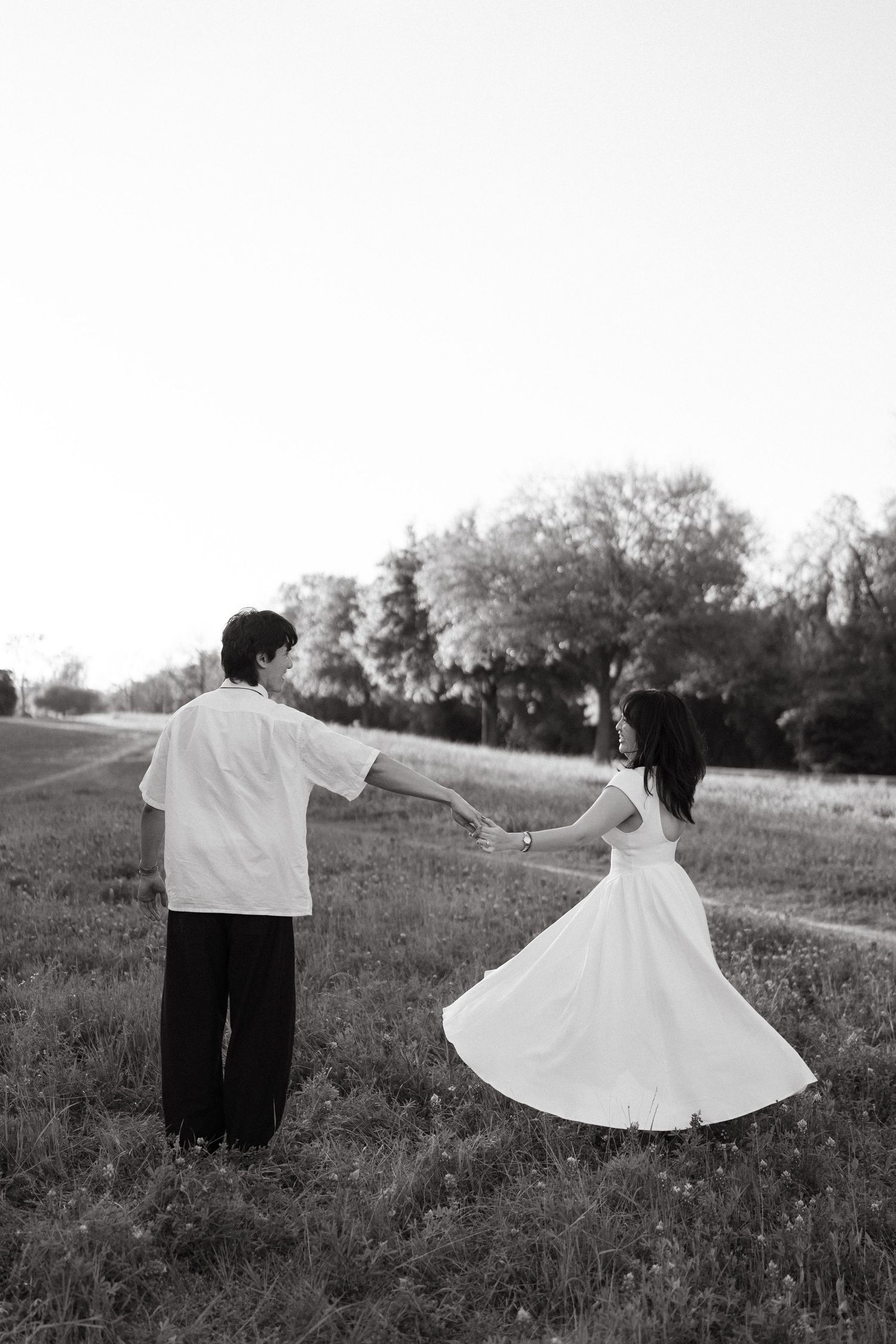 A couple in black and white stands in a meadow, with the woman twirling in a flowing white dress while the man watches.