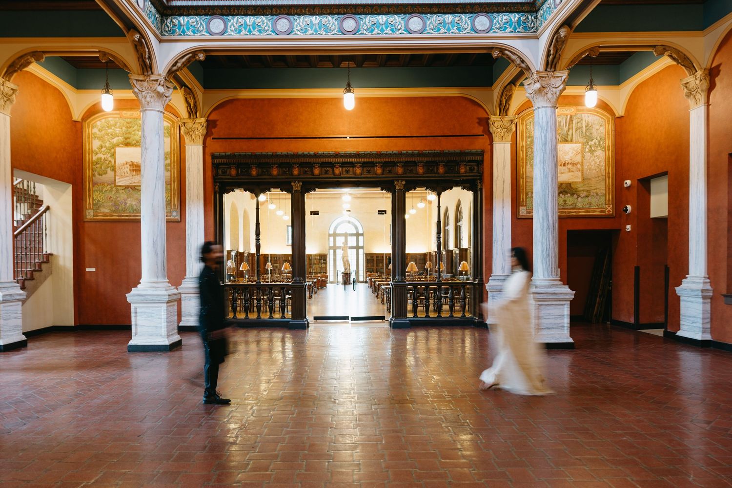 Grand historic building interior with ornate columns, murals, and elegant wooden doors, with blurred figures walking through.