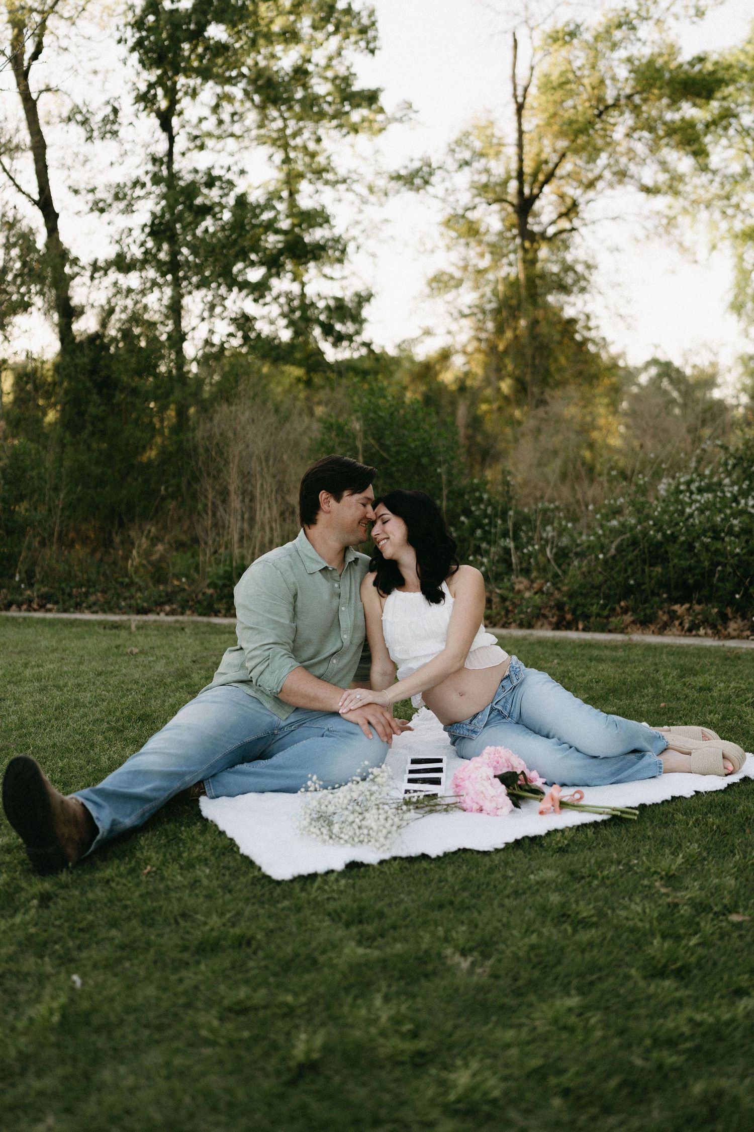 Couple sharing a romantic moment on a white blanket during a golden hour picnic in a lush green park with pink flowers.