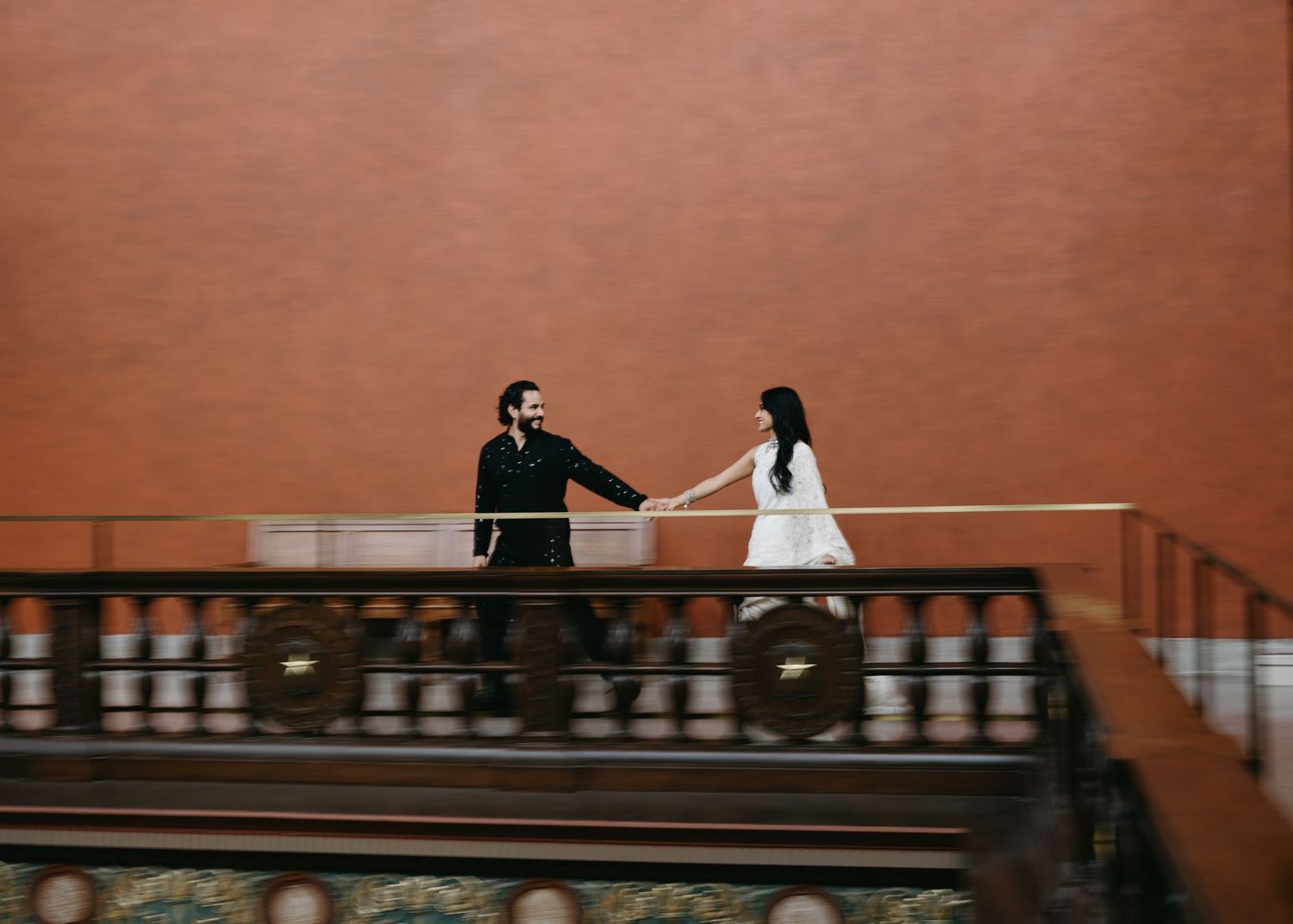 A couple holds hands on an ornate balcony railing against a warm brick wall backdrop.