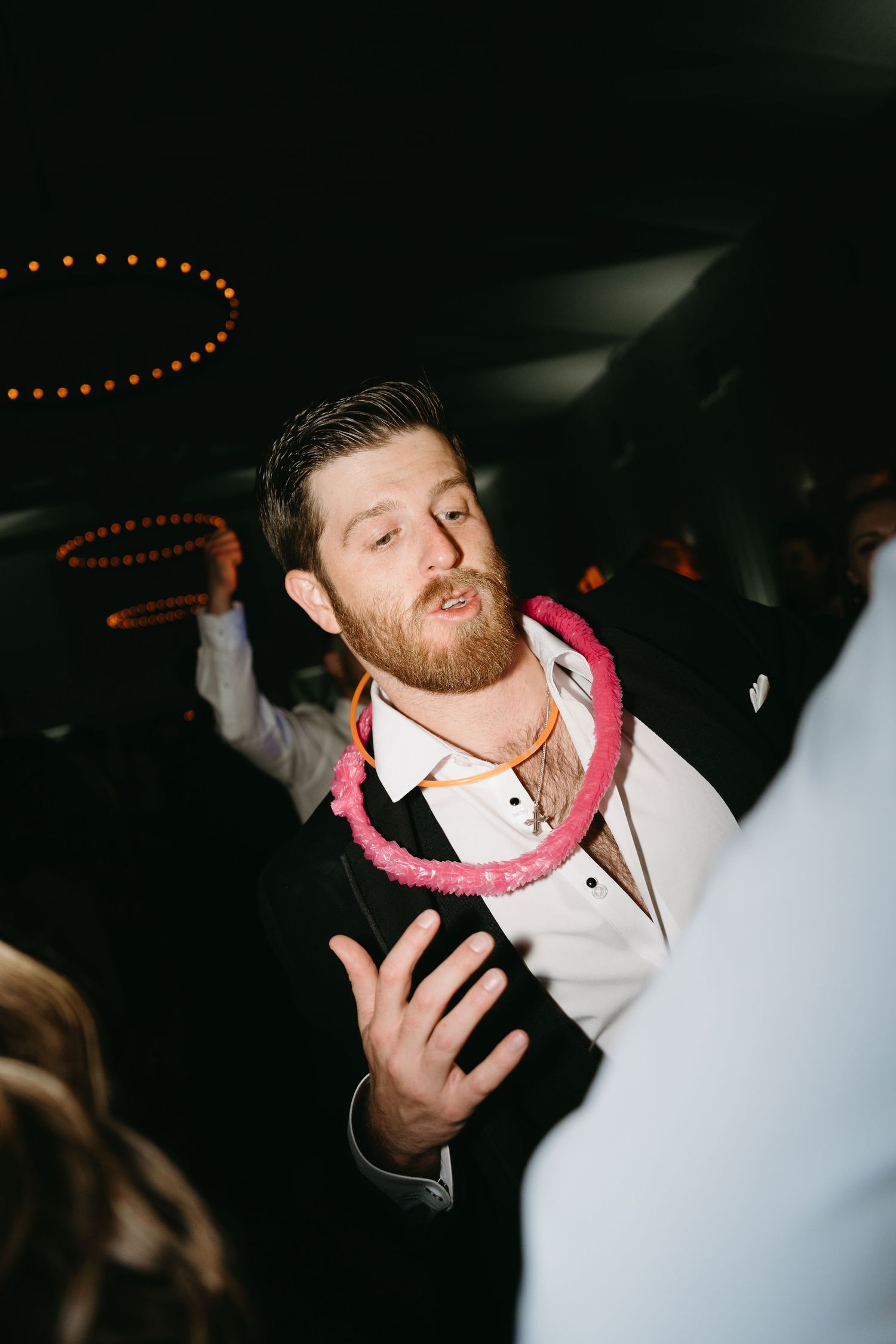 A person with a beard wearing a white shirt and pink accessory gestures expressively in a dimly lit setting.