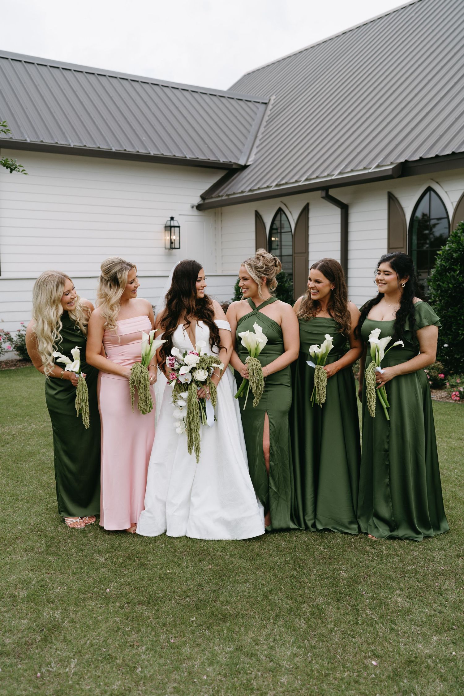 Bride in white gown with bridesmaids in pink and green dresses holding floral bouquets outside a white chapel.