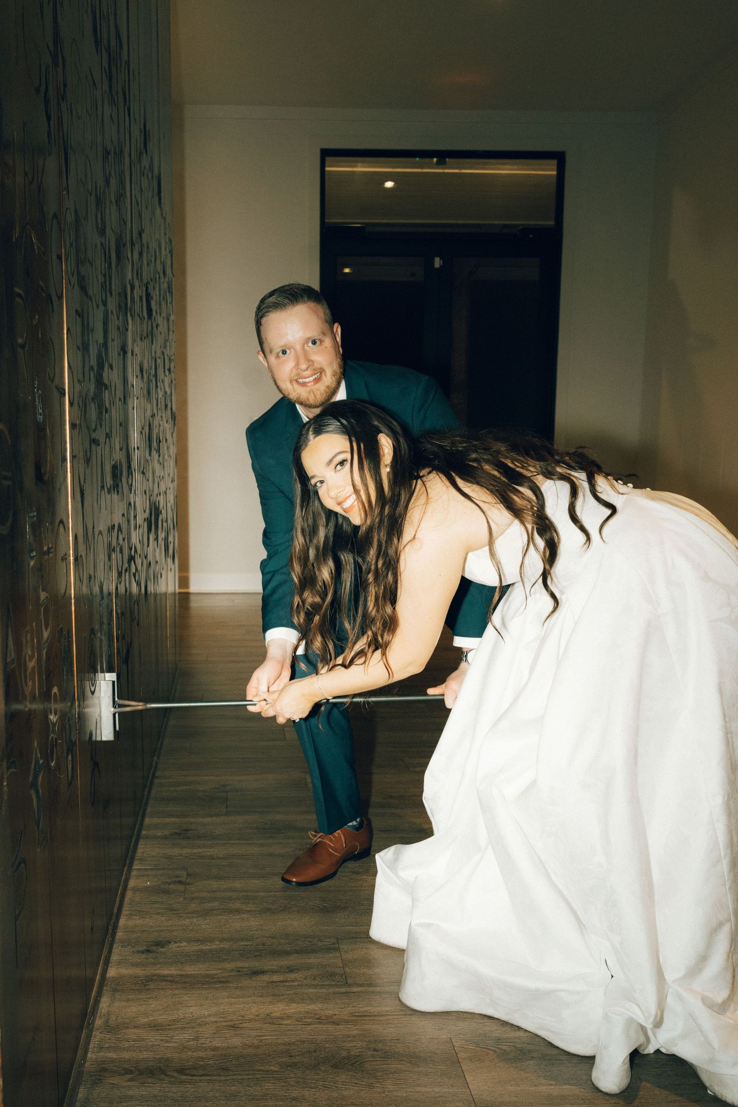Bride in white gown and groom in teal suit playfully putt golf together indoors on their wedding day.