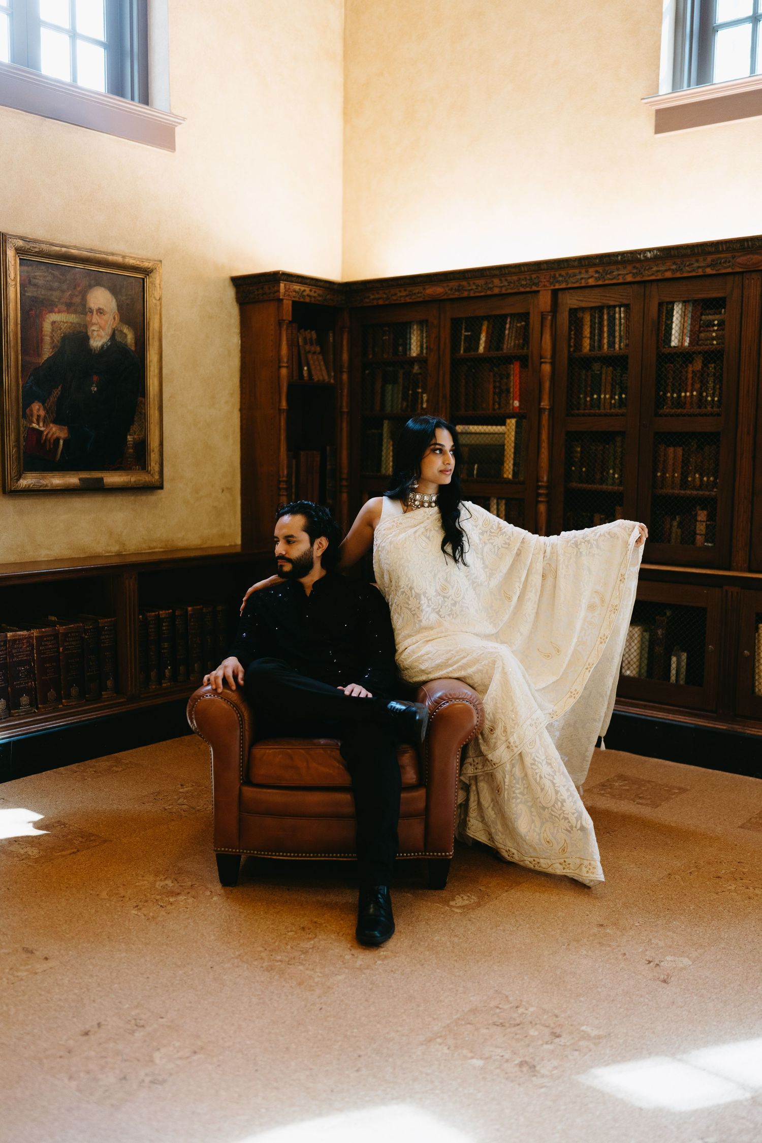 A bride in a white gown and groom in black sit together in a leather chair inside an elegant wood-paneled library.