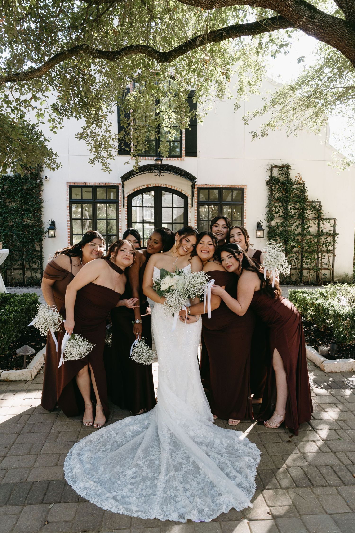 A wedding party shares candid laughing moments together on a patio with white walls and trees overhead.
