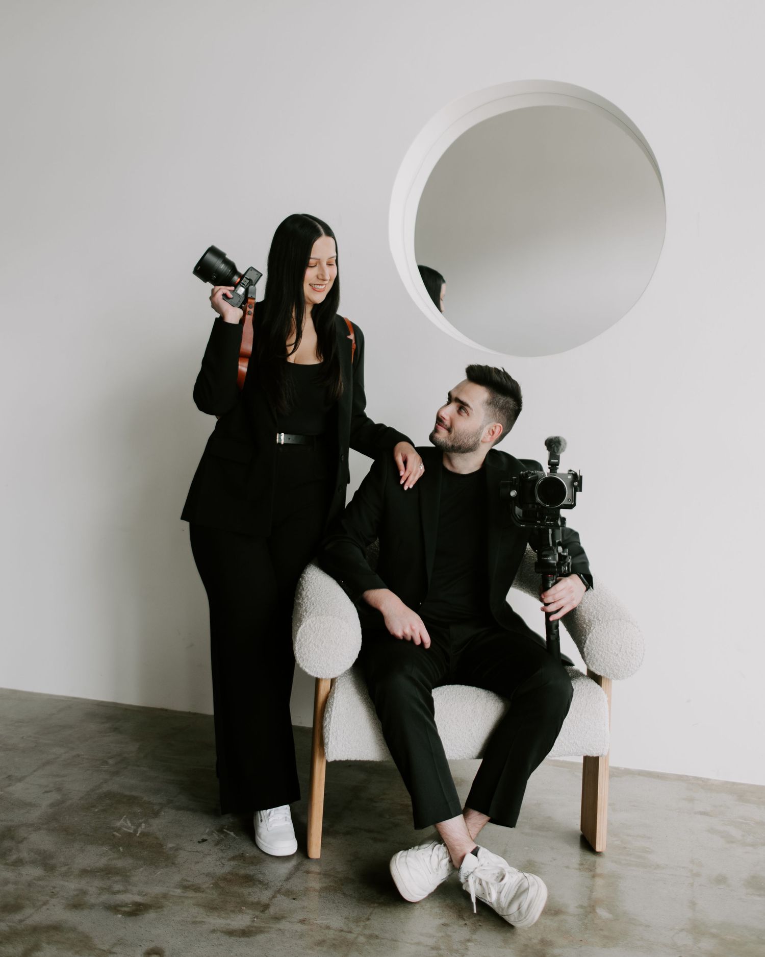 Two people in black and white outfits pose together in a minimalist studio setting with various camera equipment.