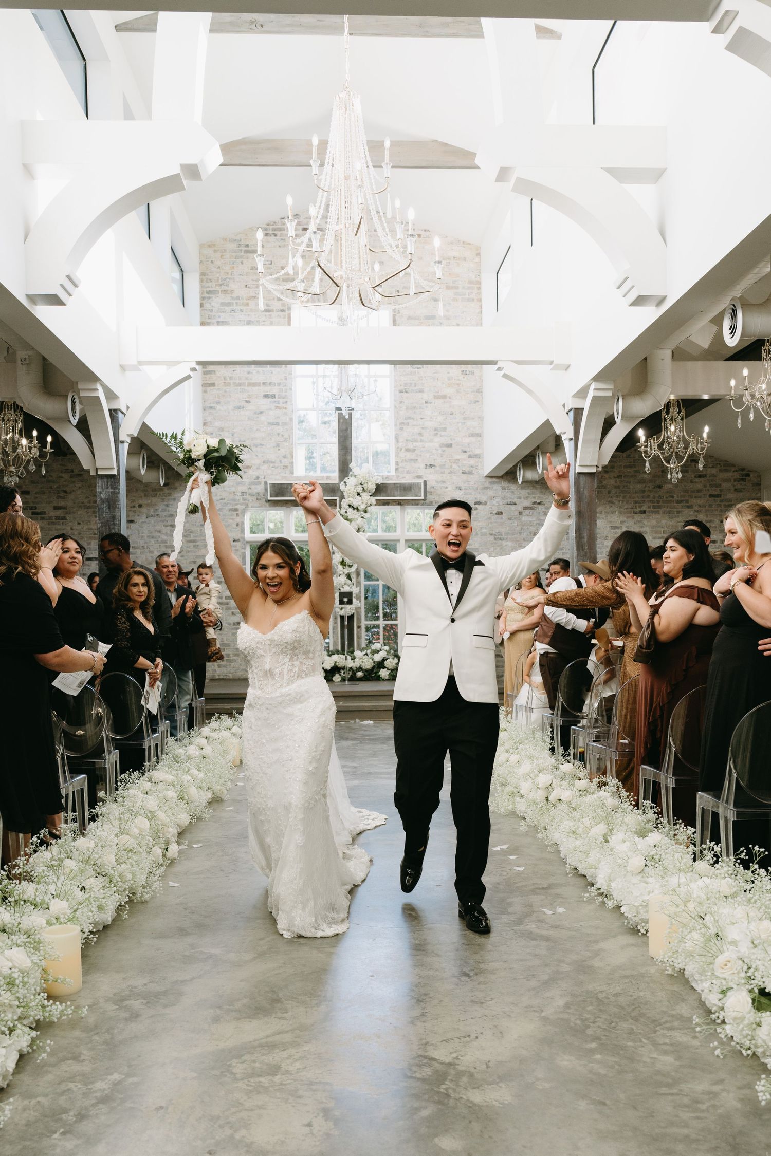 Wedding party celebrates joyfully down white floral-lined aisle in elegant venue with crystal chandeliers.