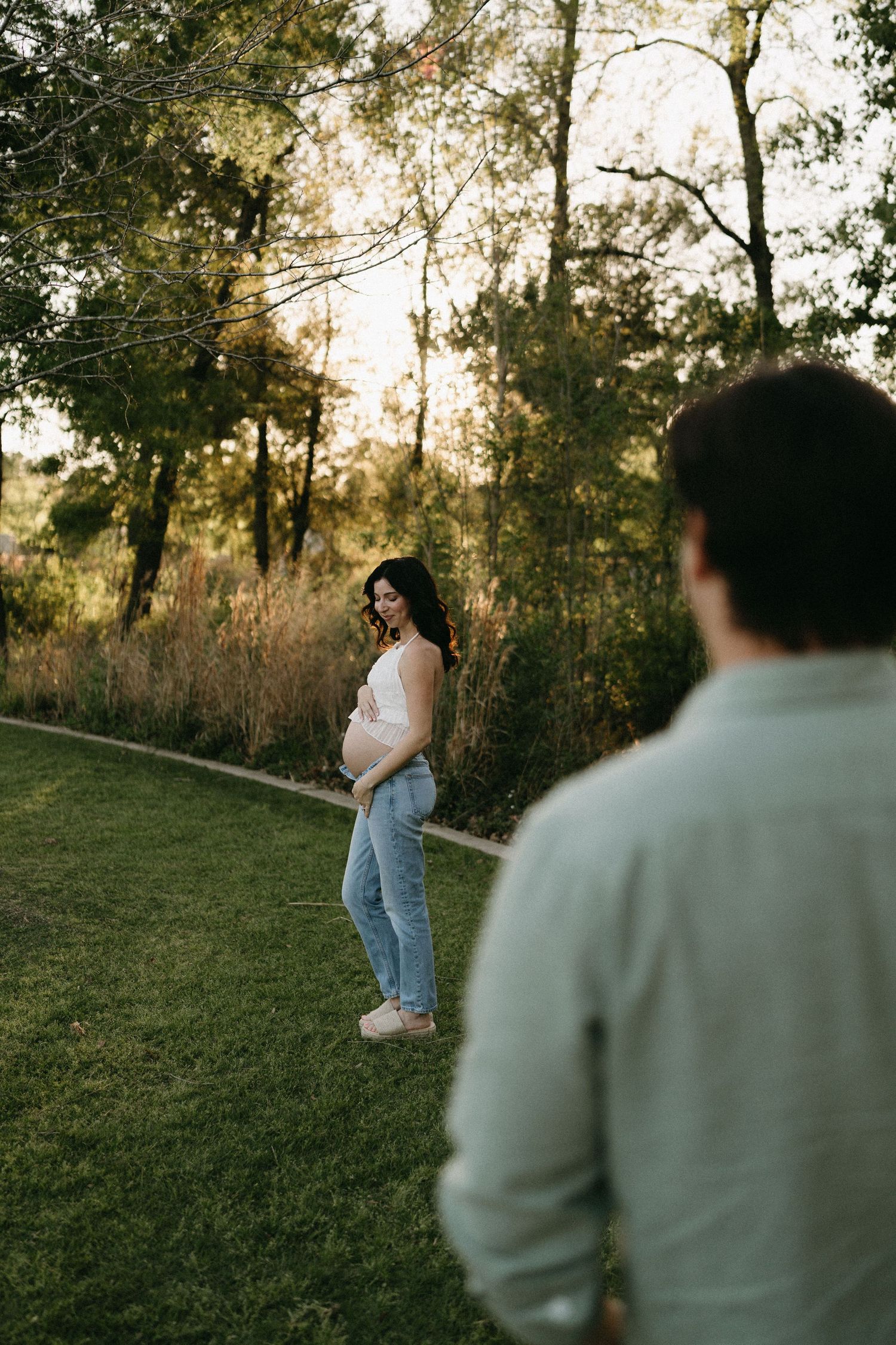 Pregnant woman in white top and jeans smiling outdoors at golden hour, with partner in foreground in a lush green park.