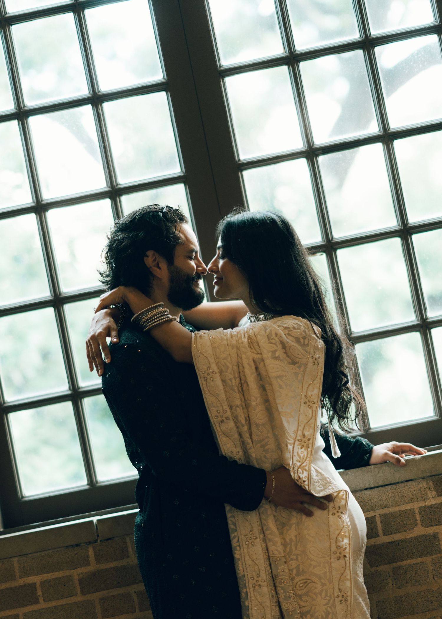 Couple in wedding attire embracing near large windows, sharing a romantic moment in soft natural light.