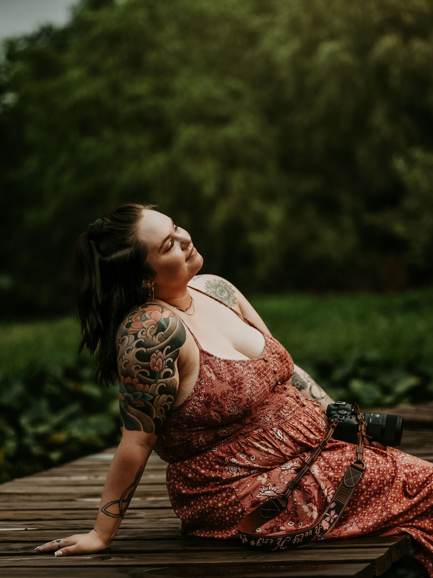 A tattooed woman in a rust-colored polka dot dress reclines gracefully on a weathered wooden dock near lush greenery.