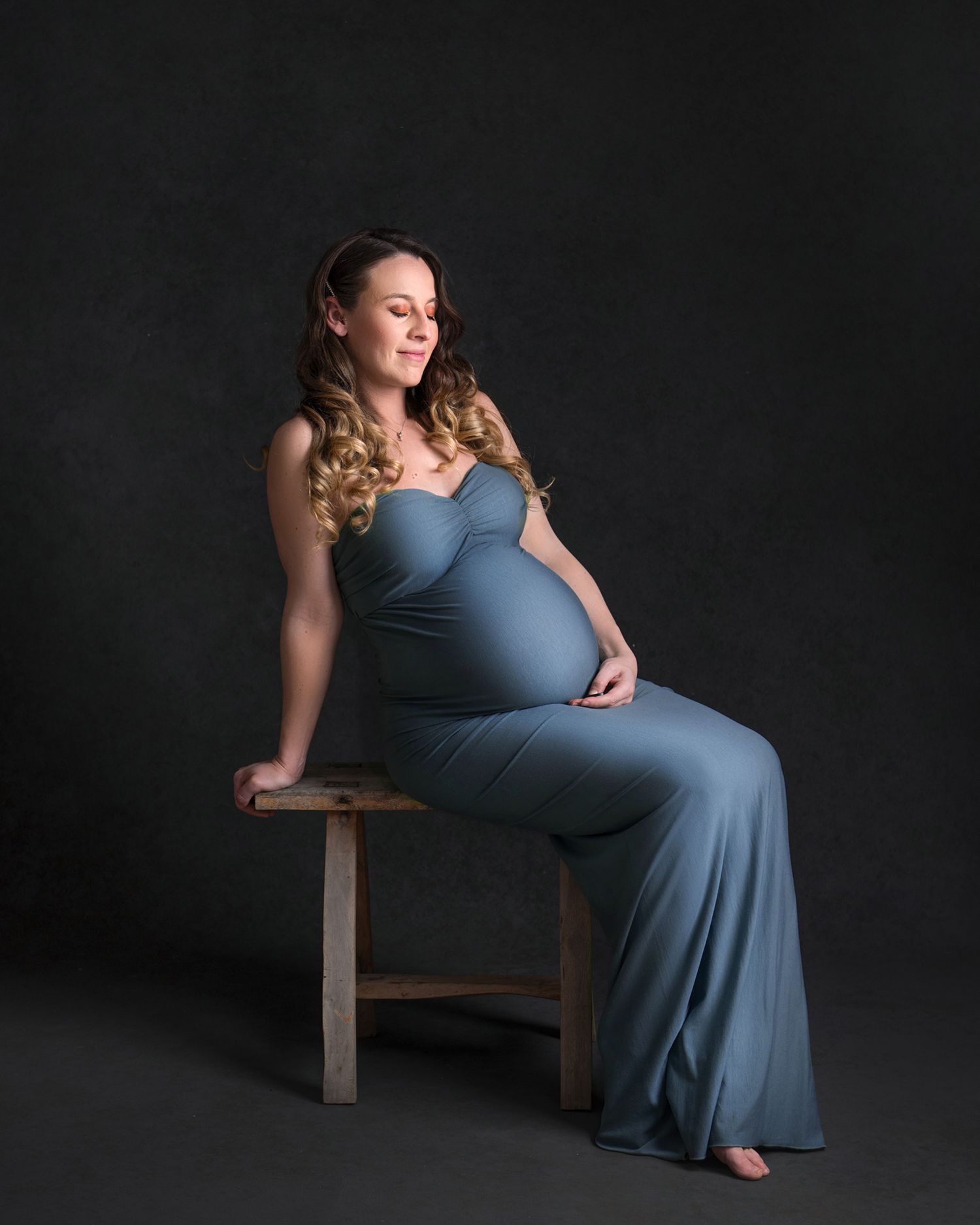 Elegant maternity portrait in blue dress against dark backdrop while seated on wooden chair.