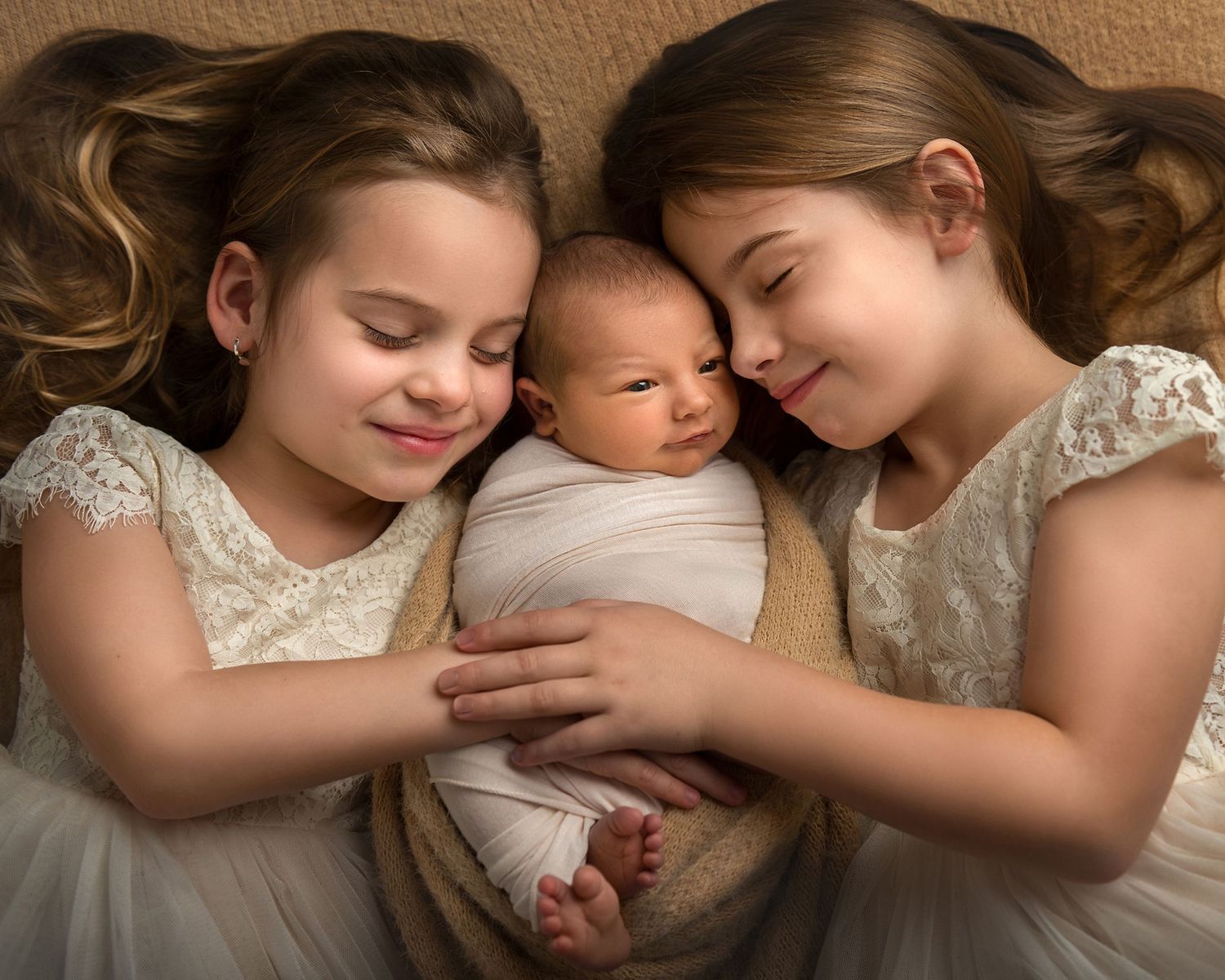 Three children in white dresses cuddle together on beige couch.